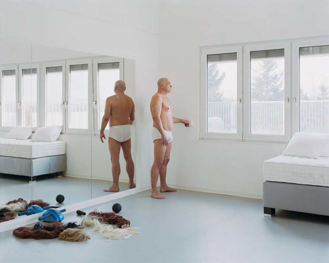 Color photograph of a man standing in his underwear in a white bedroom with various wigs on the floor