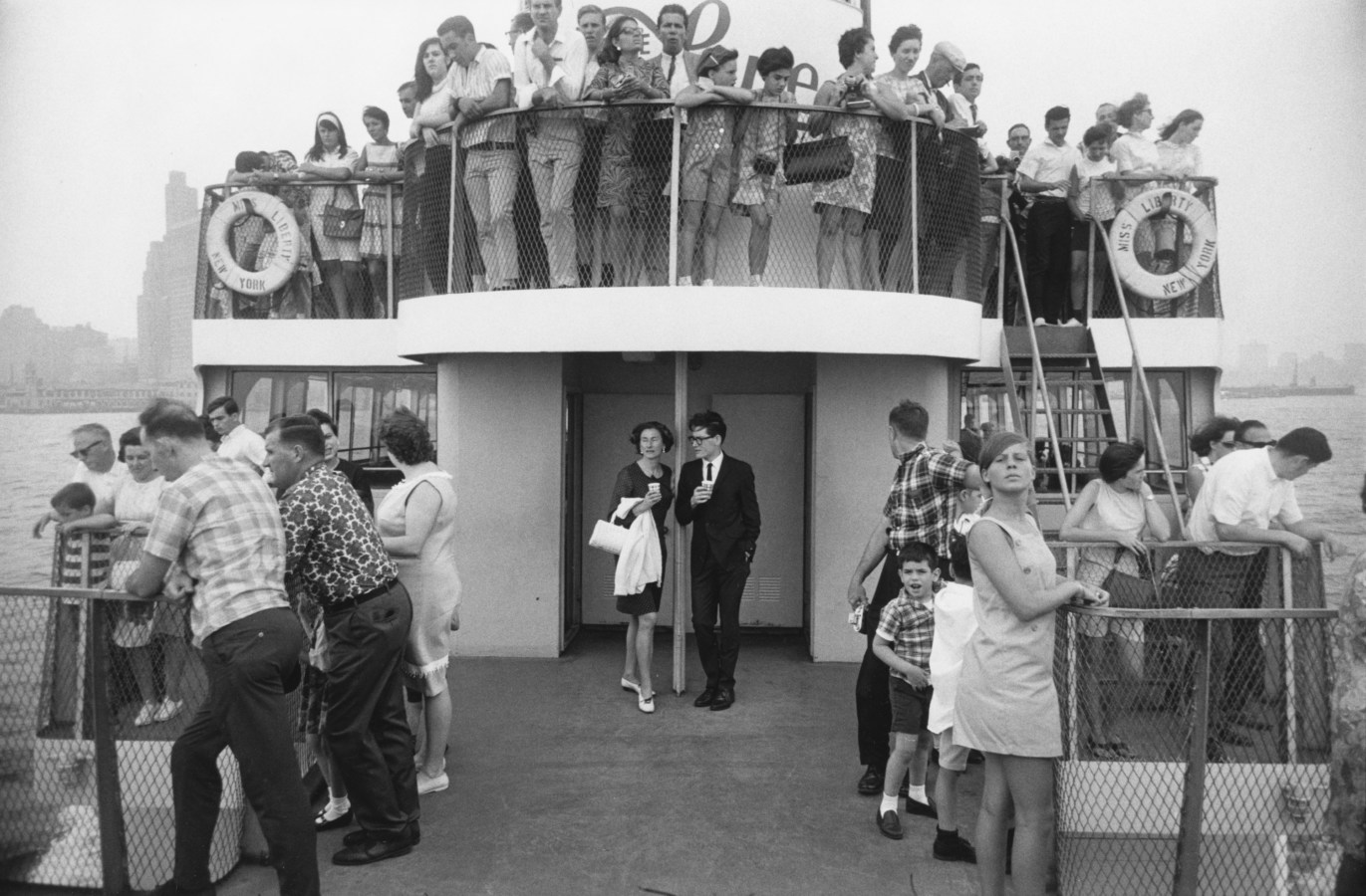 Black-and-white photograph of a young man and woman at the front of a two-story ferry filled with people looking out over the railings