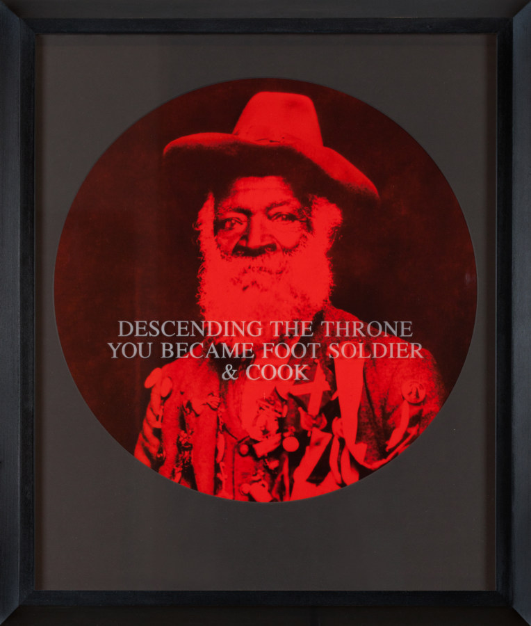 A circular photograph printed in red and black tones depicts a bearded man staring into the camera, framed in a black frame. Text printed across the image reads "Descending the Throne You Became Foot Soldier & Cook"
