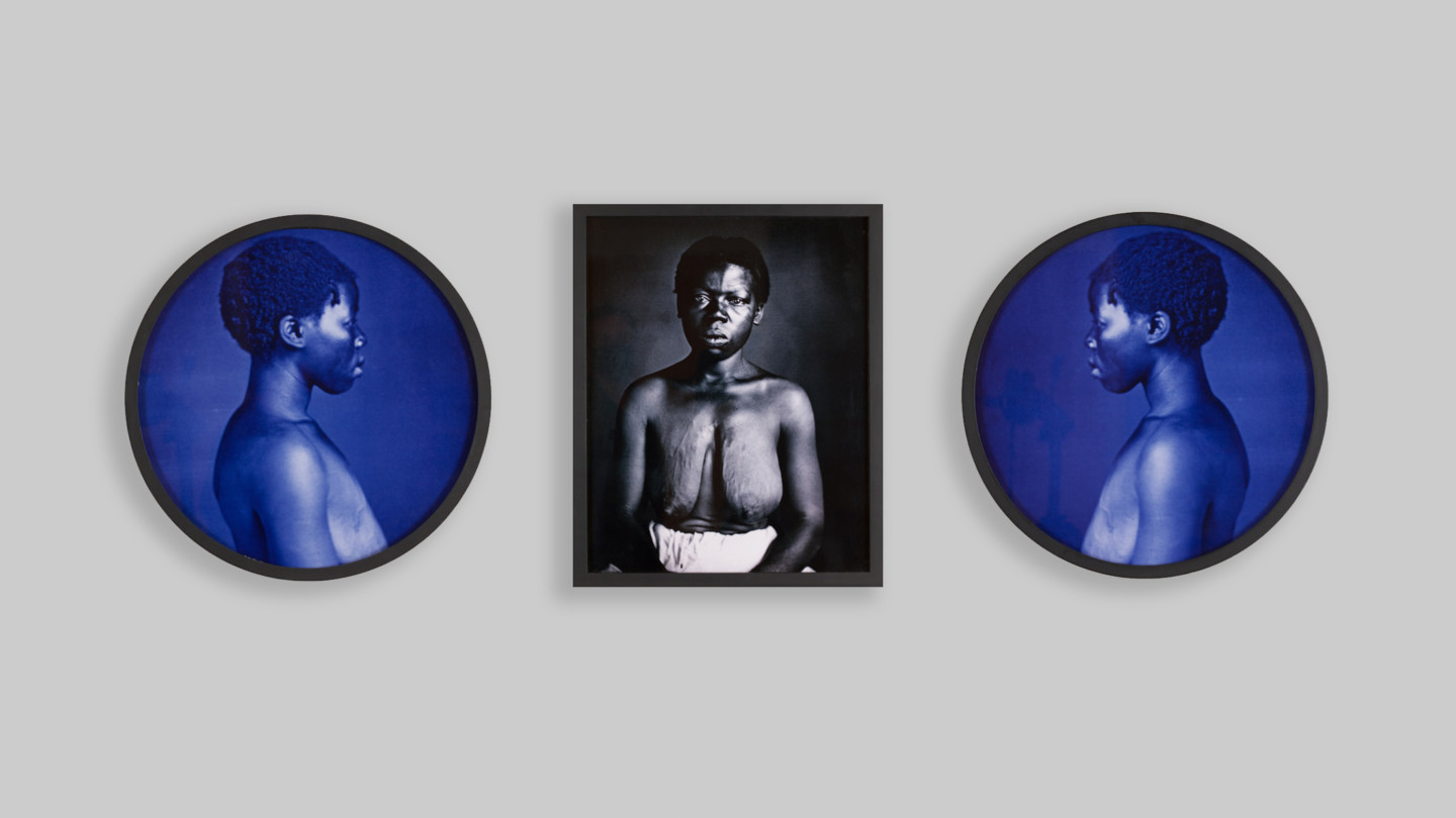 Installation view of one framed black and white portrait of an enslaved woman with her blouse removed, staring directly into the camera, accompanied by two framed blue and black portraits of the same woman looking down in profile.