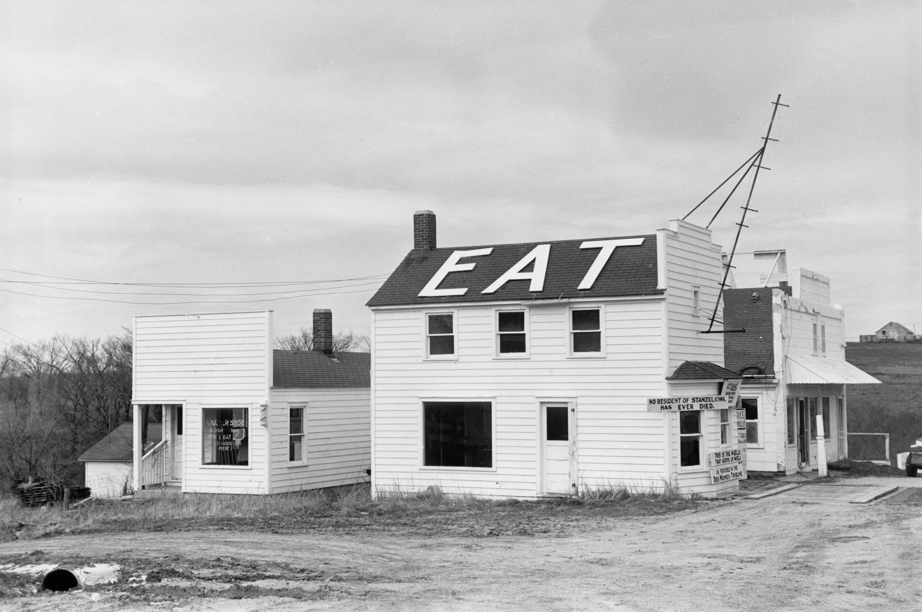 Black and white photograph of a rural two story building with the word 'eat' in large white letters on its roof.