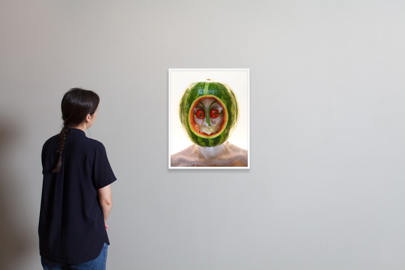 Installation photograph of a person observing a framed color photograph of a person with a watermelon over their head.