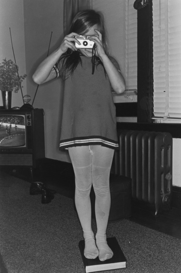Black and white photograph of a young girl taking a photograph in a living room
