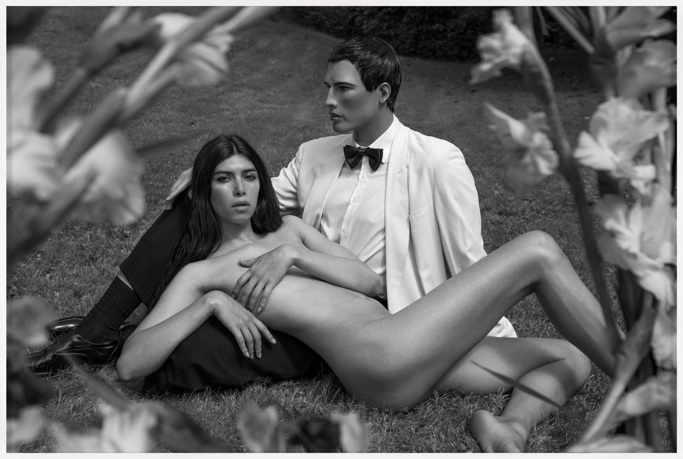 Black and white photograph of a reclining nude woman leaning against a male mannequin in a suit