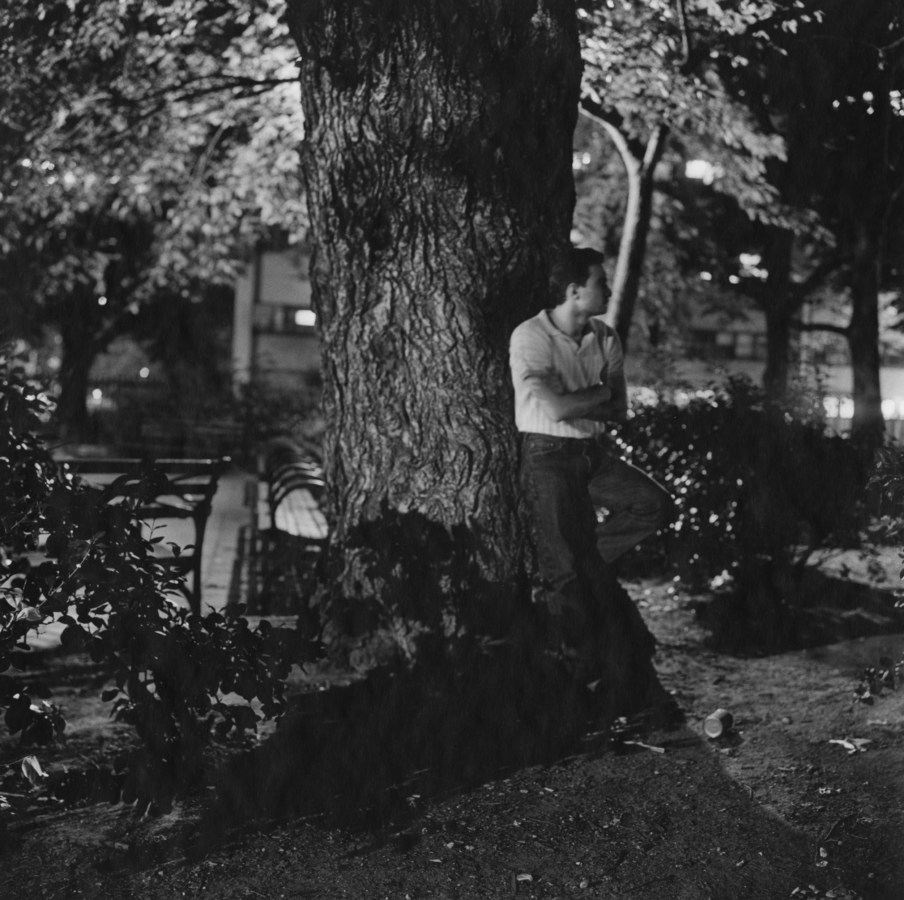 Black and white photograph of a man leaning against a tree in a park at night