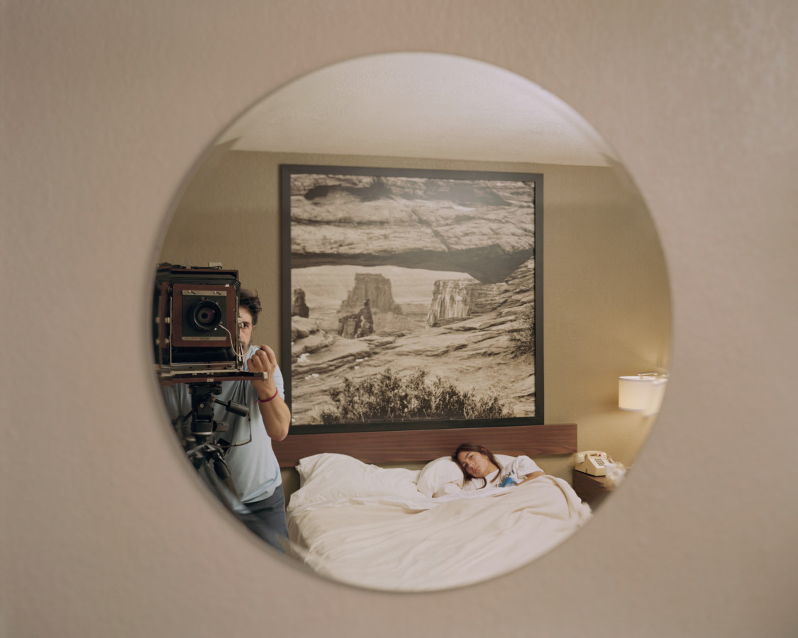 Color photograph of a round mirror reflecting the photographer standing next to a woman lying in bed