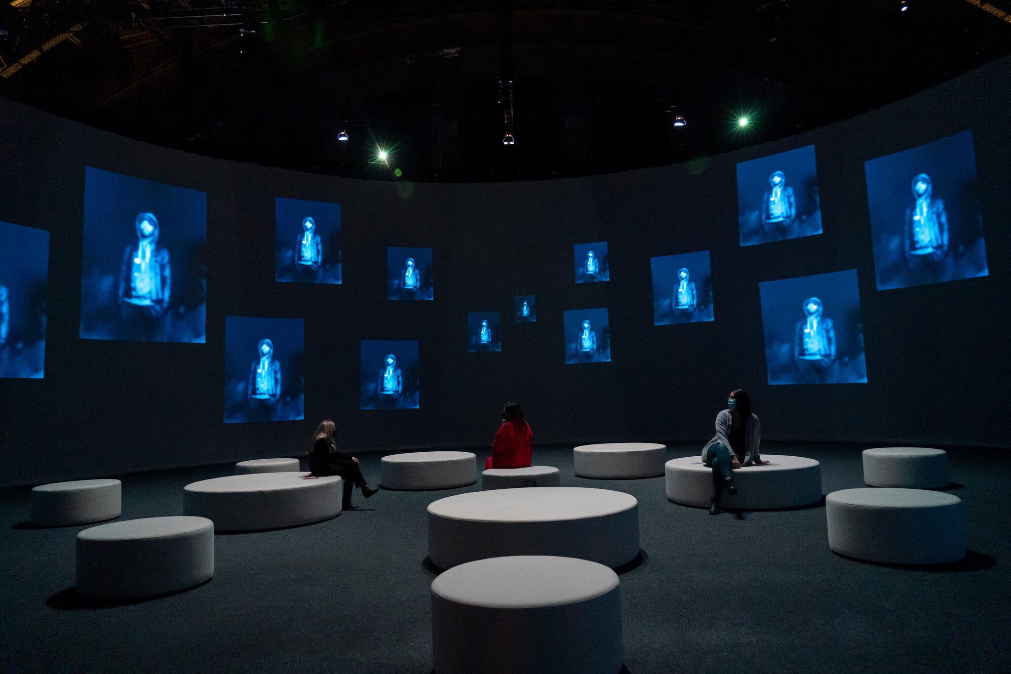 Installation photograph of people seated in a gallery surrounded by variou screens projecting identical images of a young man wearing a hoodie