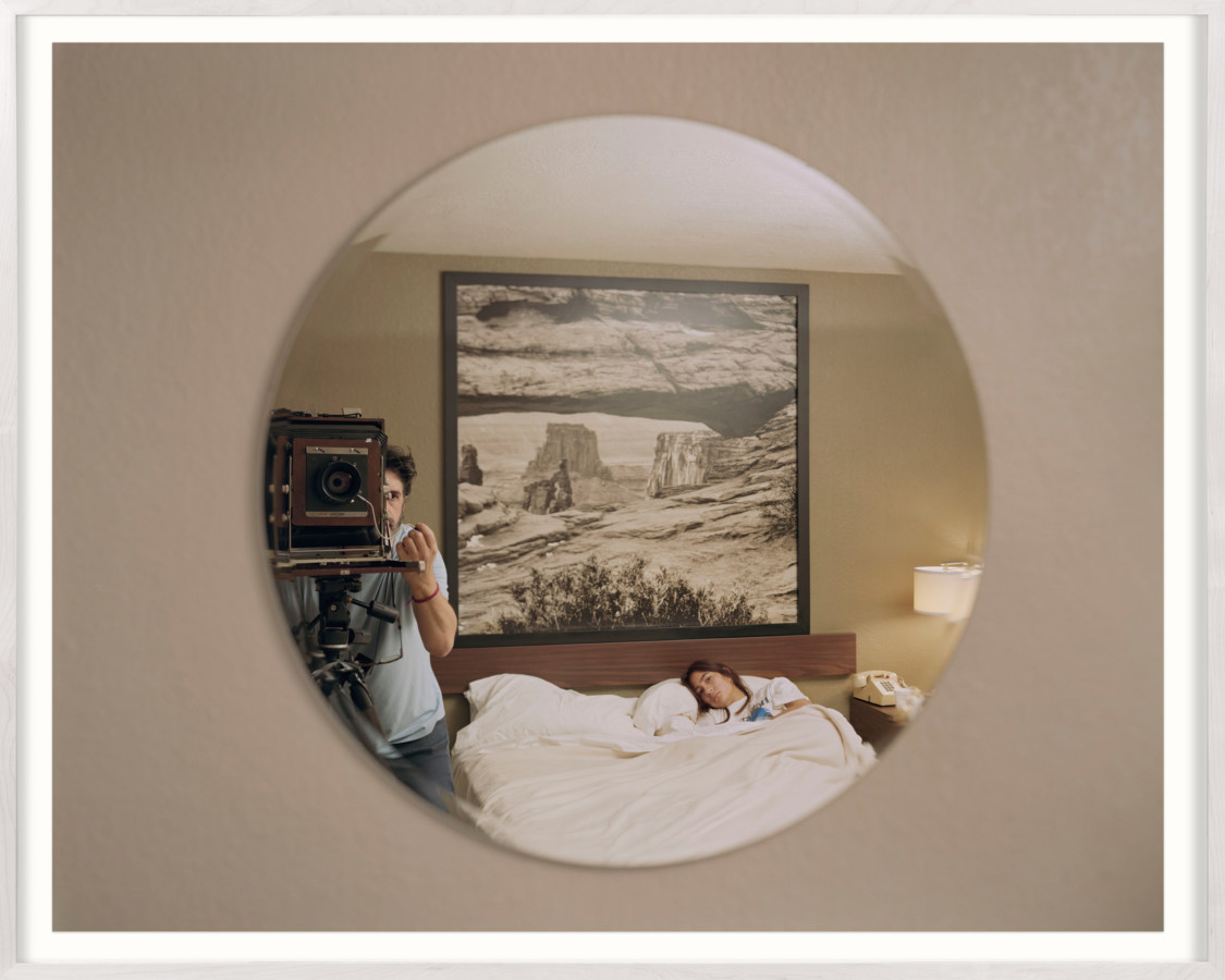 Framed color photograph of a photographer using a large format camera to take a portrait of a girl in a hotel bed through a mirror