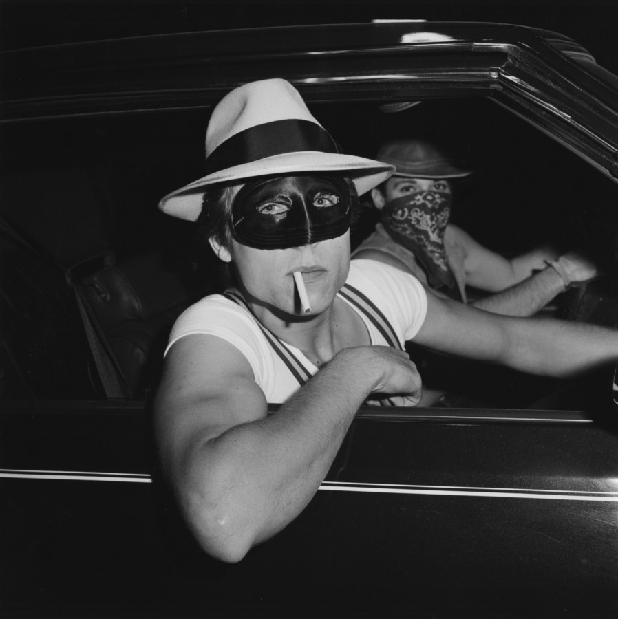 A black and white photograph of two people in a car wearing hats and masks, one leans out of the window with a cigarette in their mouth, the other peers in the same direction from the steering wheel