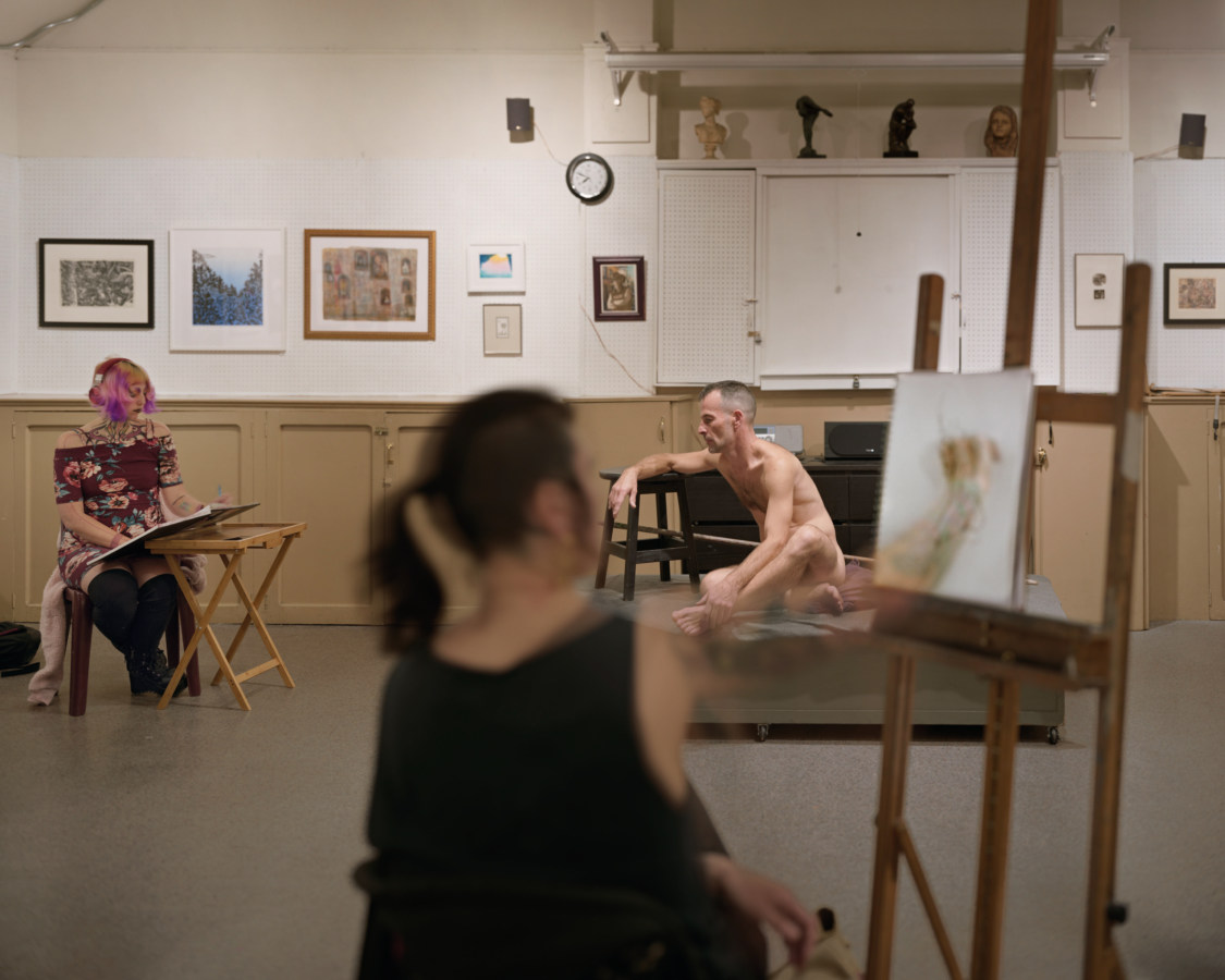 Color image of nude male model posing in life drawing classroom with two students drawing