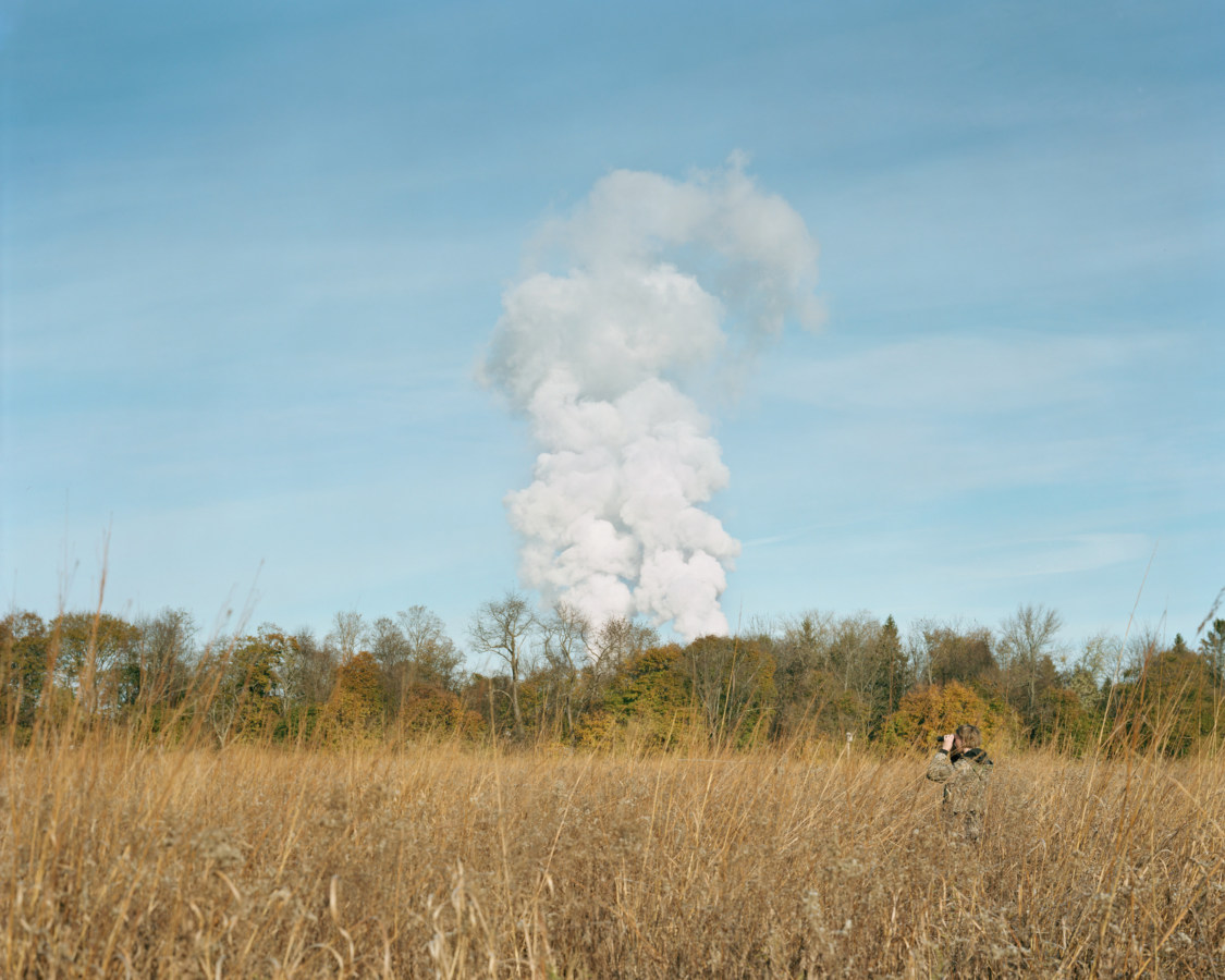 Color image of a person with binoculars in grain field with large cloud of smoke on the horizon