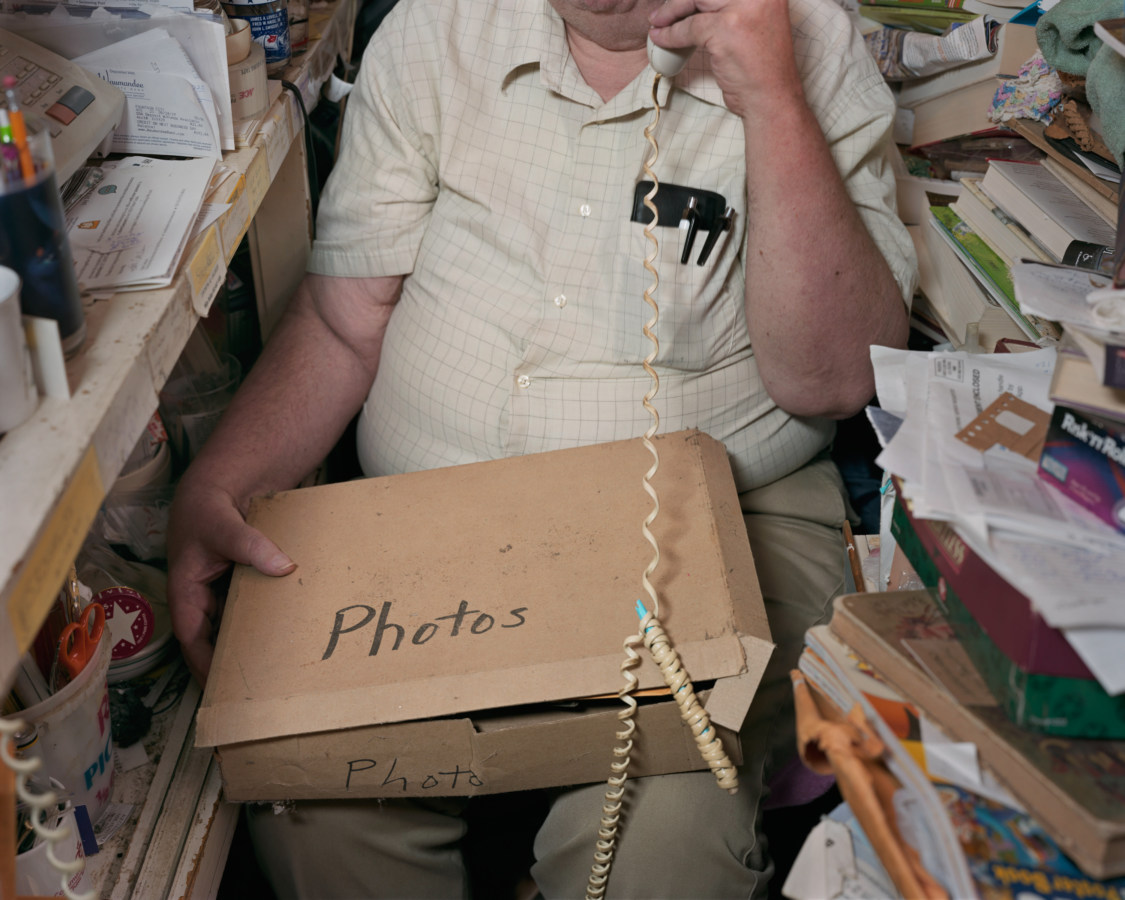 Color image of seated man holding box labeled 'Photos' surrounded by stacks of paper