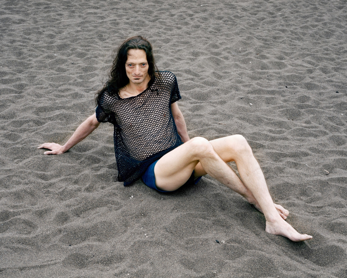 Color photograph of a long-haired man in a mesh tanktop and briefs posing on a sandy beach.