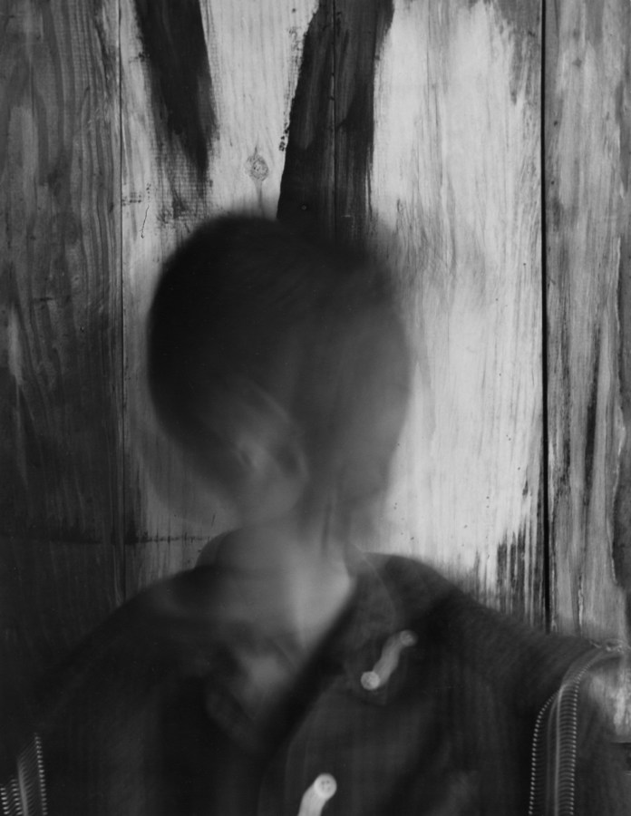 Black and white photograph of motion blurred individual in front of wood paneled wall