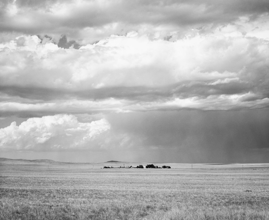 Black and white photograph of open grasslands with a small cluster of trees and houses in the distance, under a cloudy sky.
