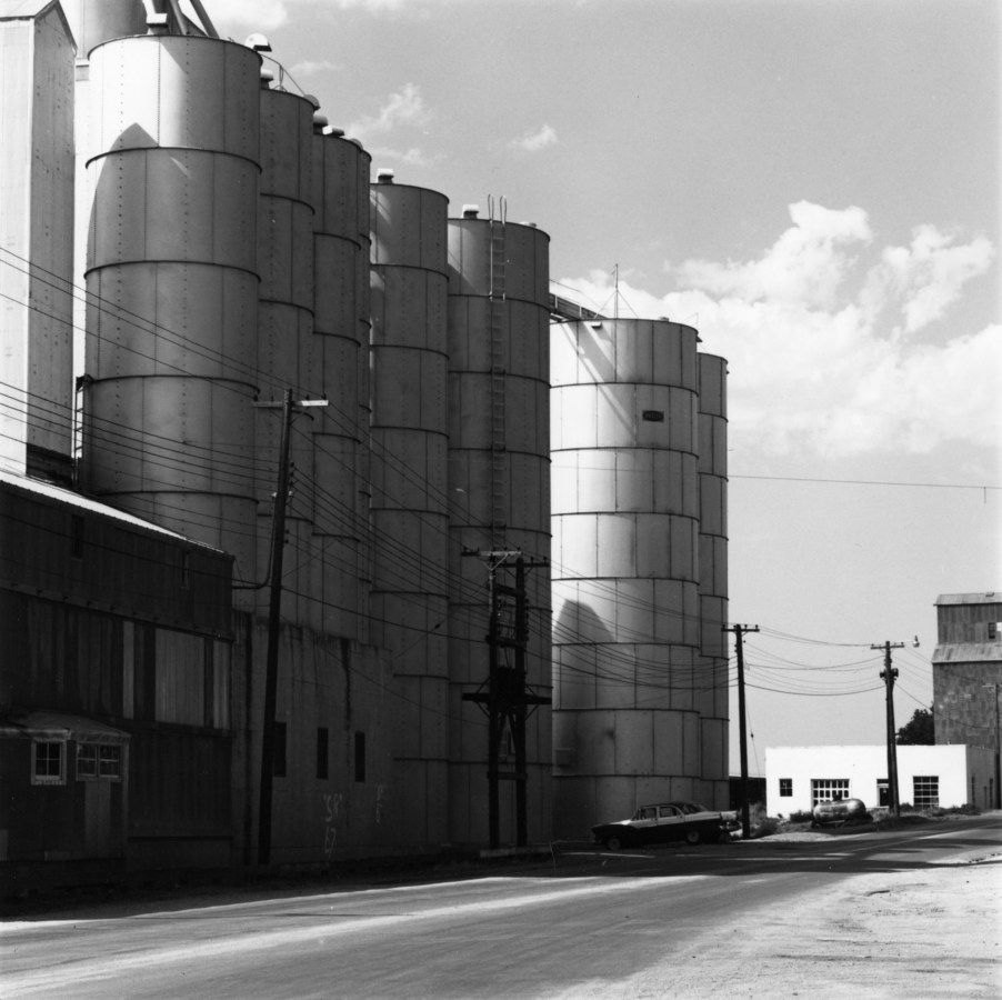 Black and white photograph of grain elevators, flanked on the right by an empty road.