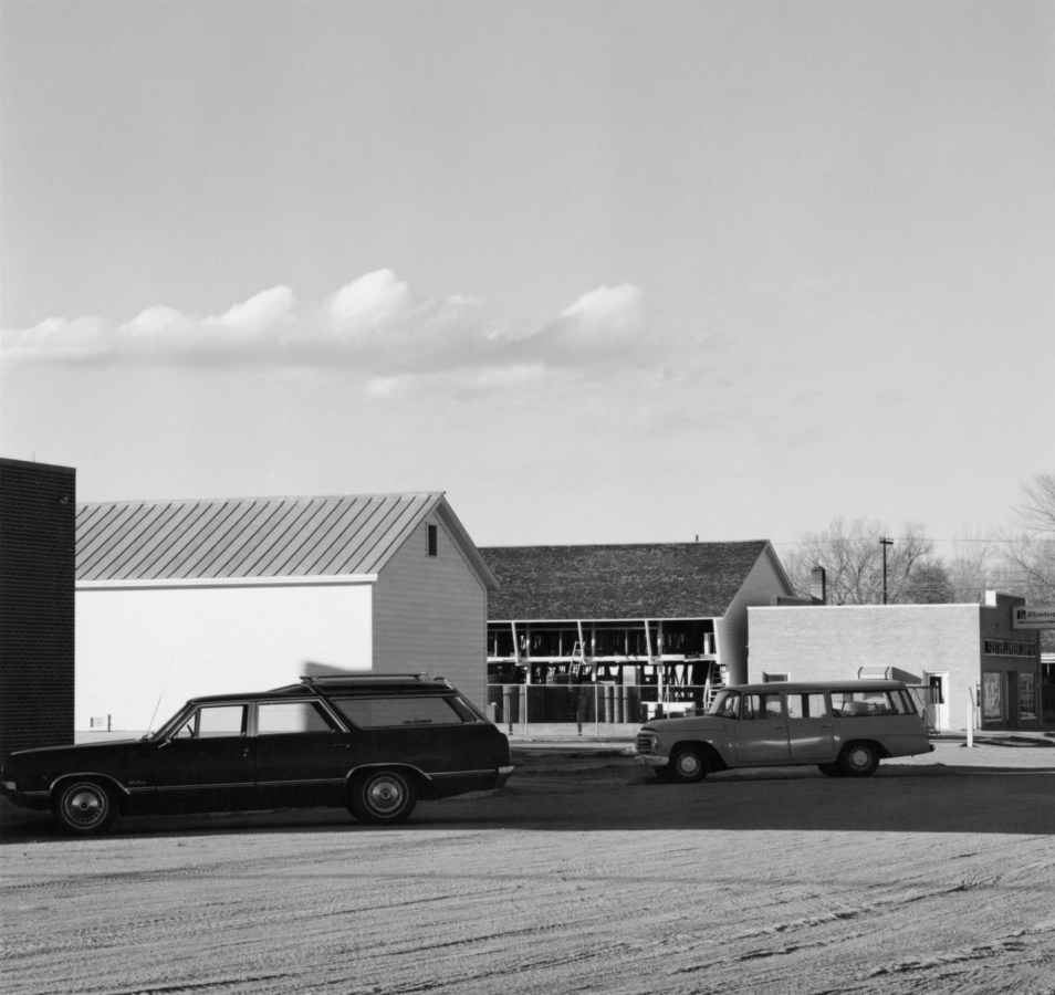 Black and white photograph of two station wagons parked in front of buildings on a dirt road.