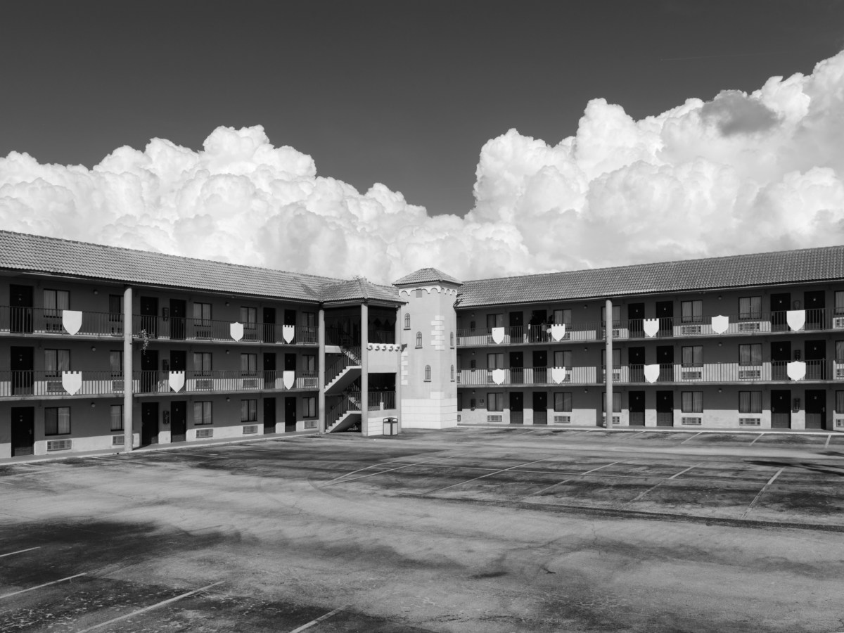 Black and white photograph of motel parking lot with looming clouds just over the building