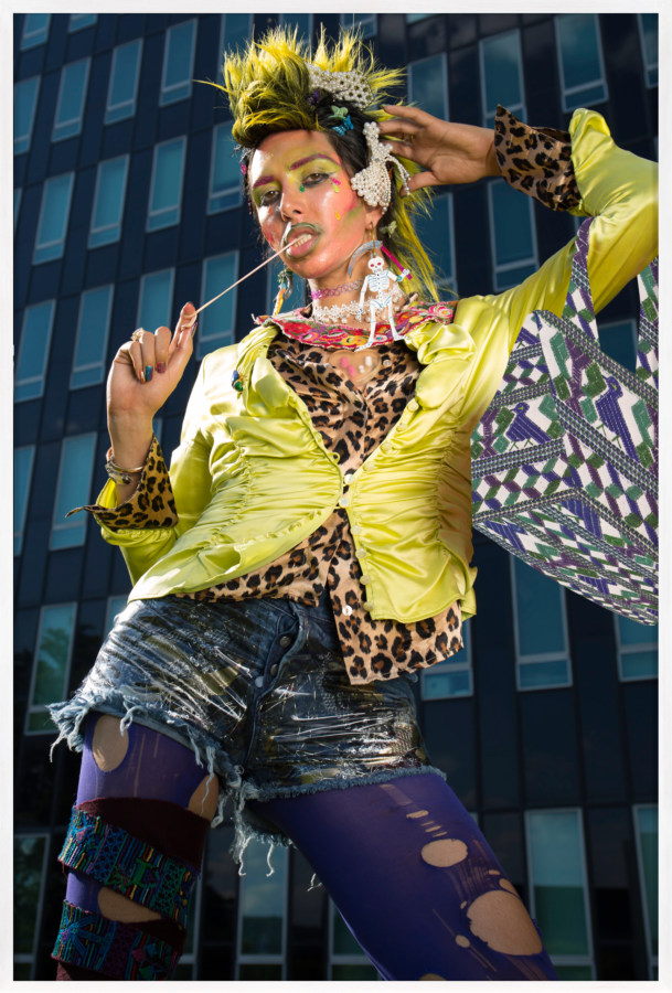 Color photograph of a model dressed in vibrant colors and loud patterns chewing gum in front of building framed in white