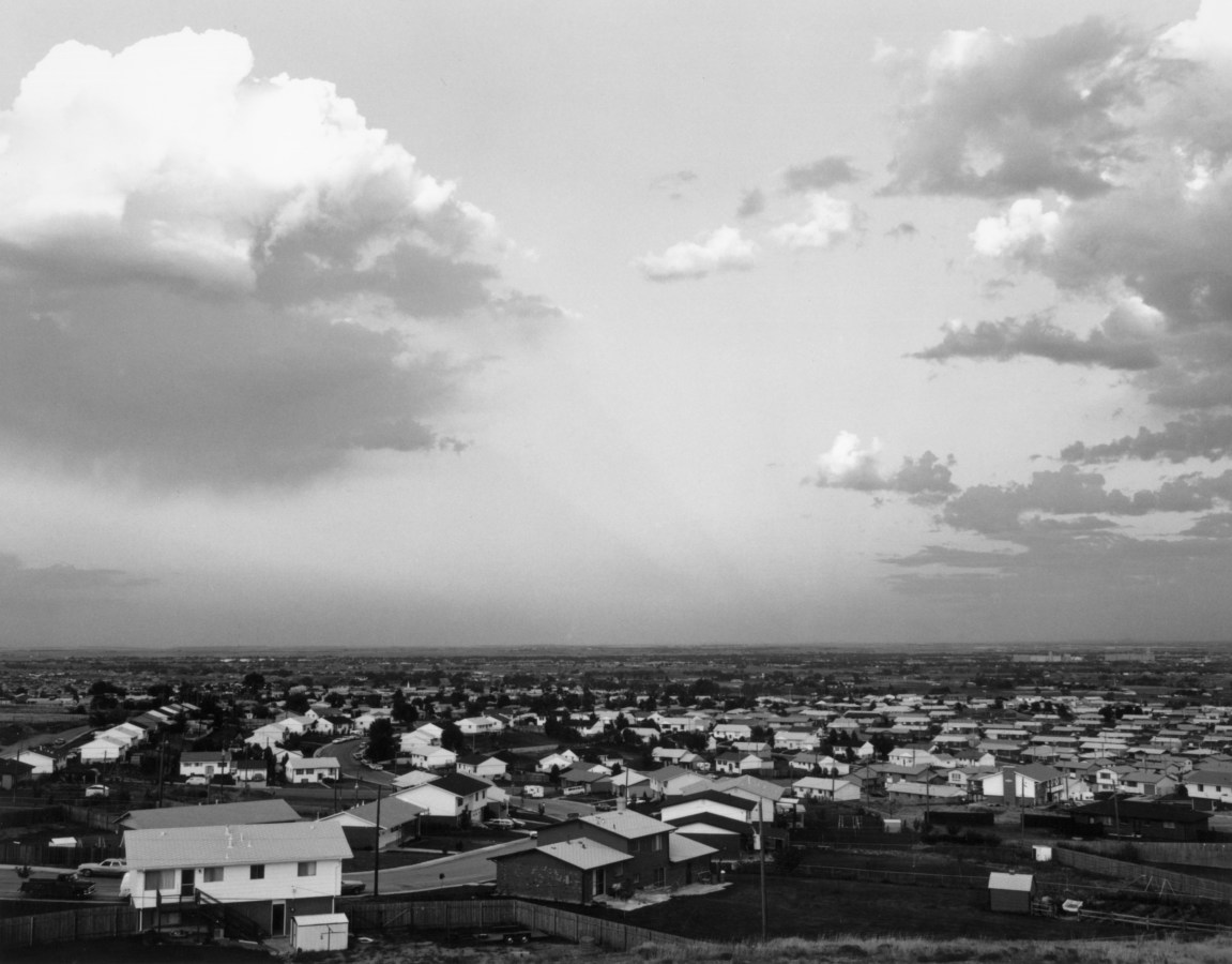 A black and white image depicting a suburban neighborhood seen from an elevated area, with a flat horizon line and cloudy sky
