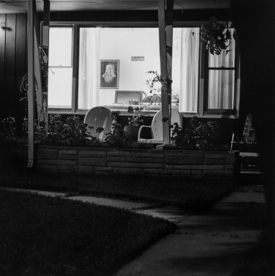 Black and white photograph of the front of a suburban home at night, with two chairs on the front porch and the home interior visible through the window.