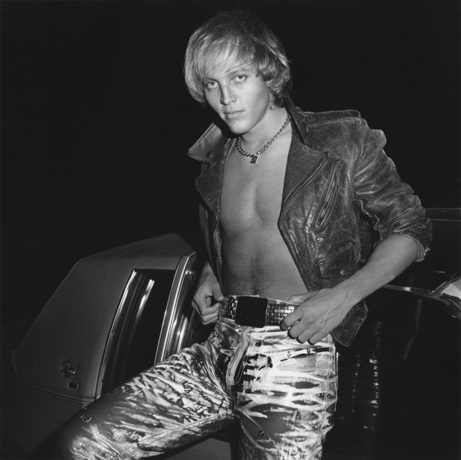 Black and white photograph of shirtless man dressed in flashy clothes sitting on car