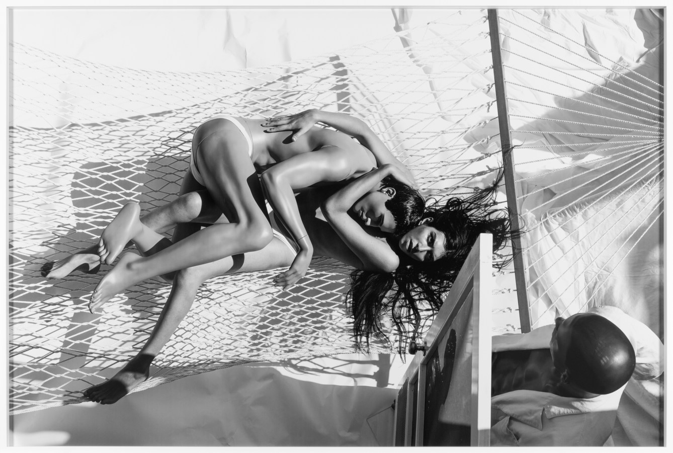 Black and white photograph of two people embracing on a hammock while another enters the space framed in white
