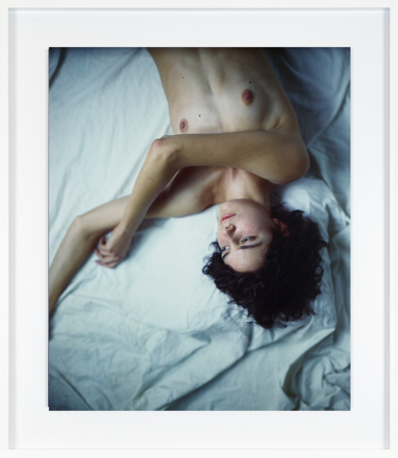 Color photograph of a nude woman laying on sheets framed in white
