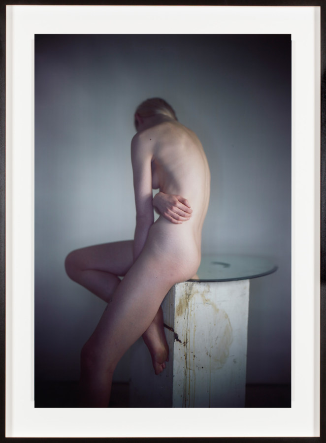 Color photograph of a seated nude figure turning away from camera framed in black