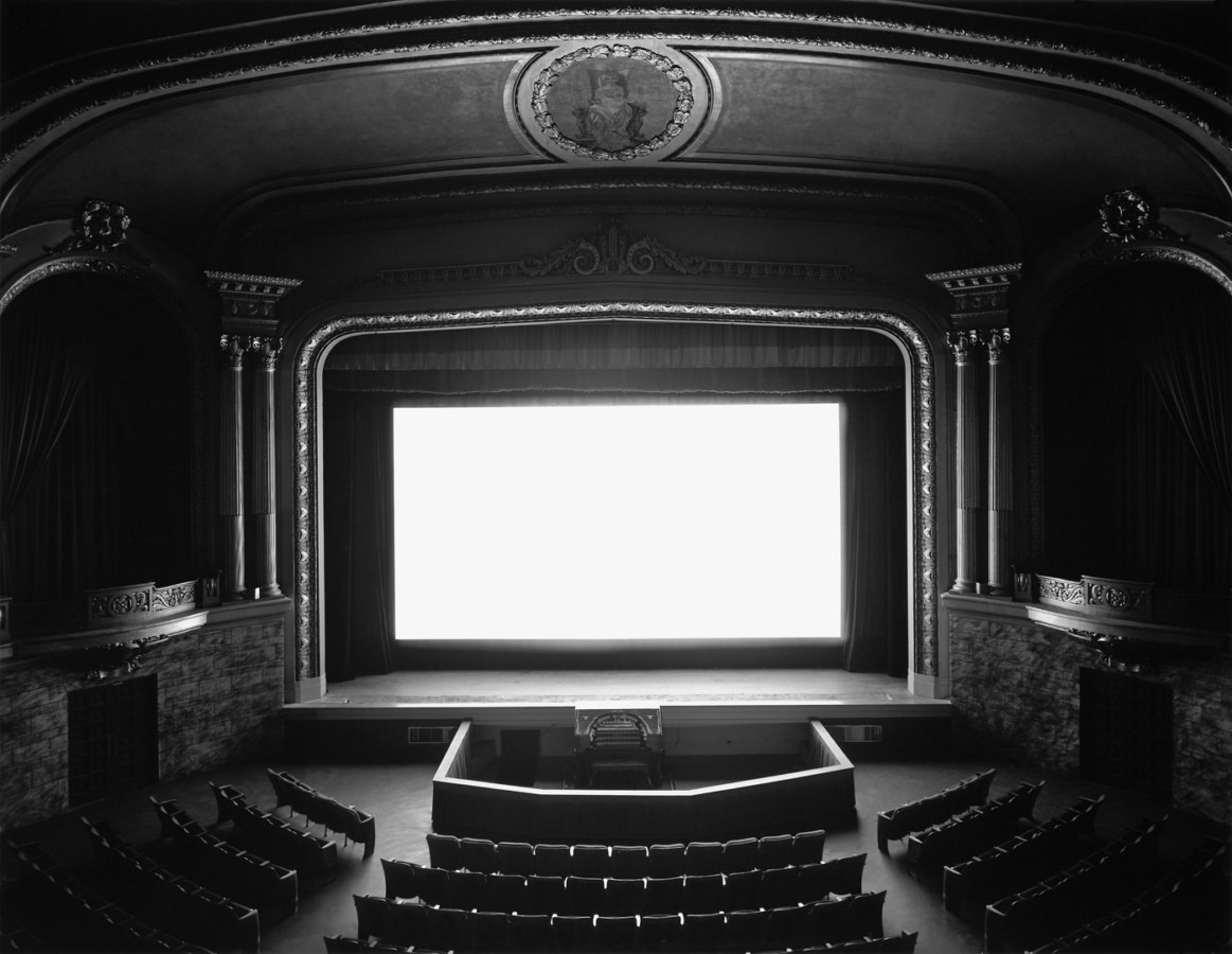 Black and white photograph of theater interior lit by screen