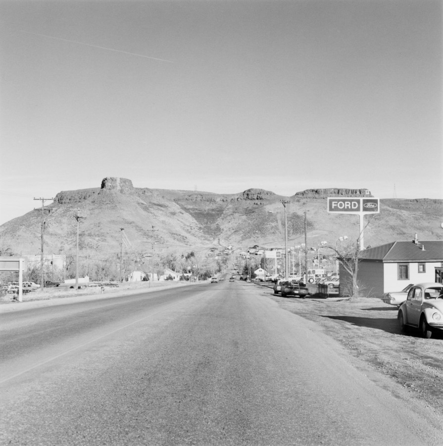 Black and white photograph of a stretch of road with businesses and homes leading to the mountains