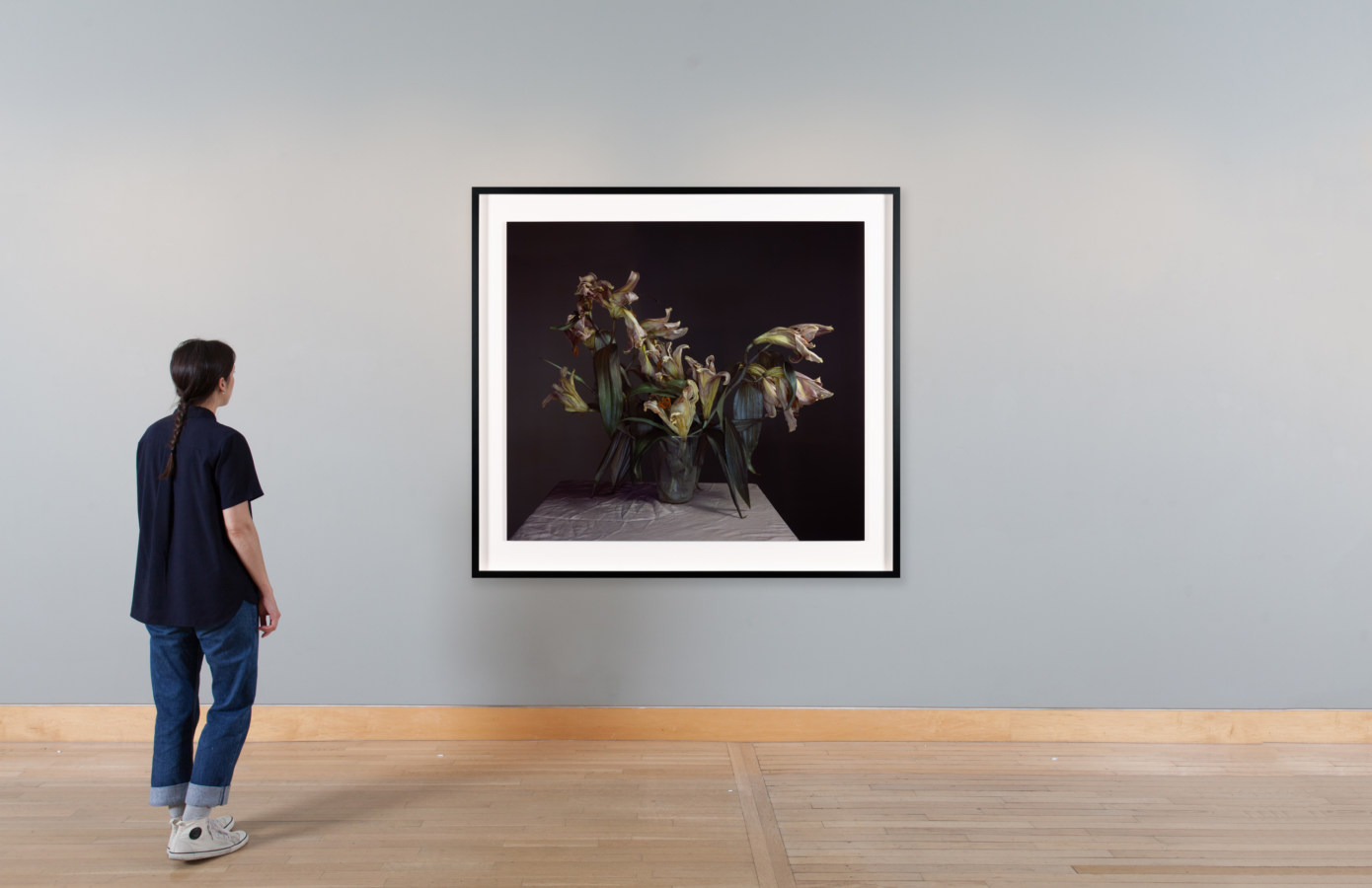 Color image of color photograph depicting decaying lilies on pink plinth framed in black on a grey gallery wall