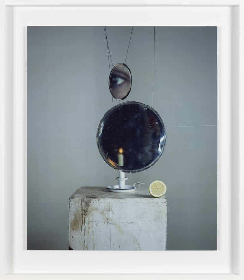 Color photograph of a candle and lemon on plinth with suspended glass and mirror reflecting an eye framed in white
