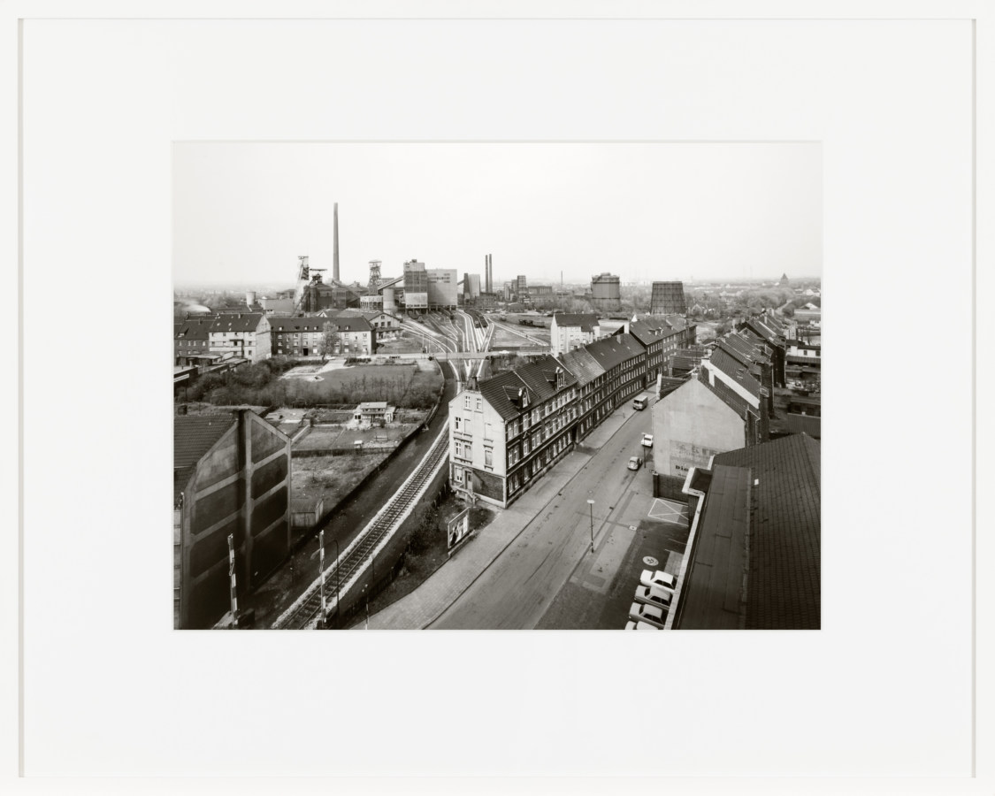 Black and white photograph of a town strip with industrial plant on the horizon framed in white