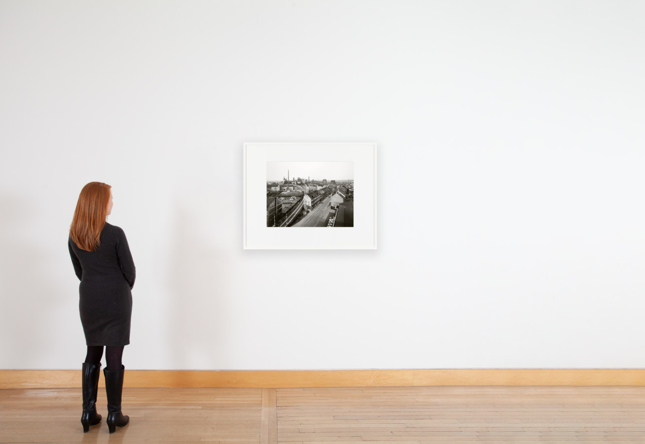 Color image of a black and white photograph depicting an industrial plant along a city framed in white and on a white gallery wall