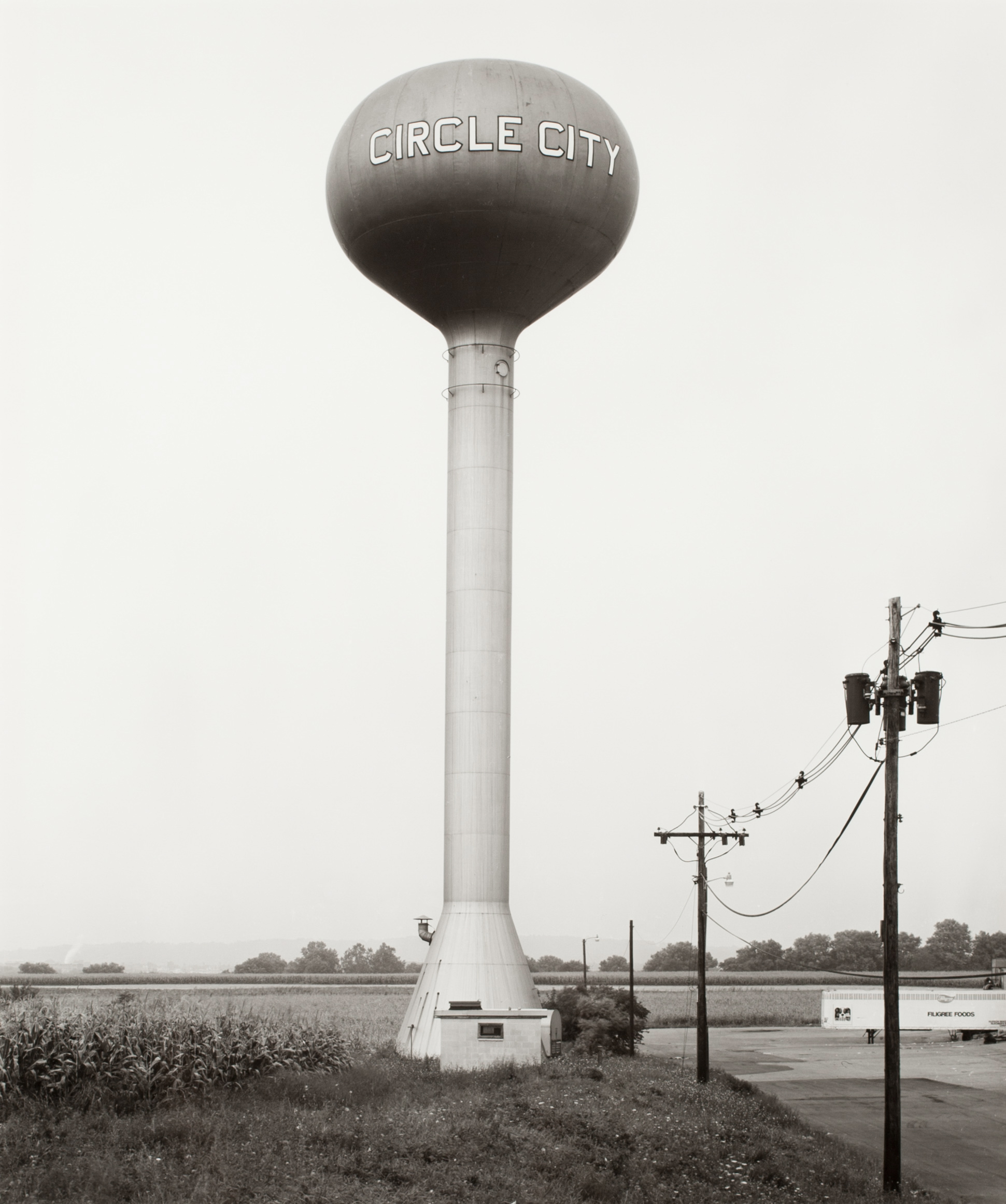 Black and white photograph of a water tower in a grass field near electrical power lines