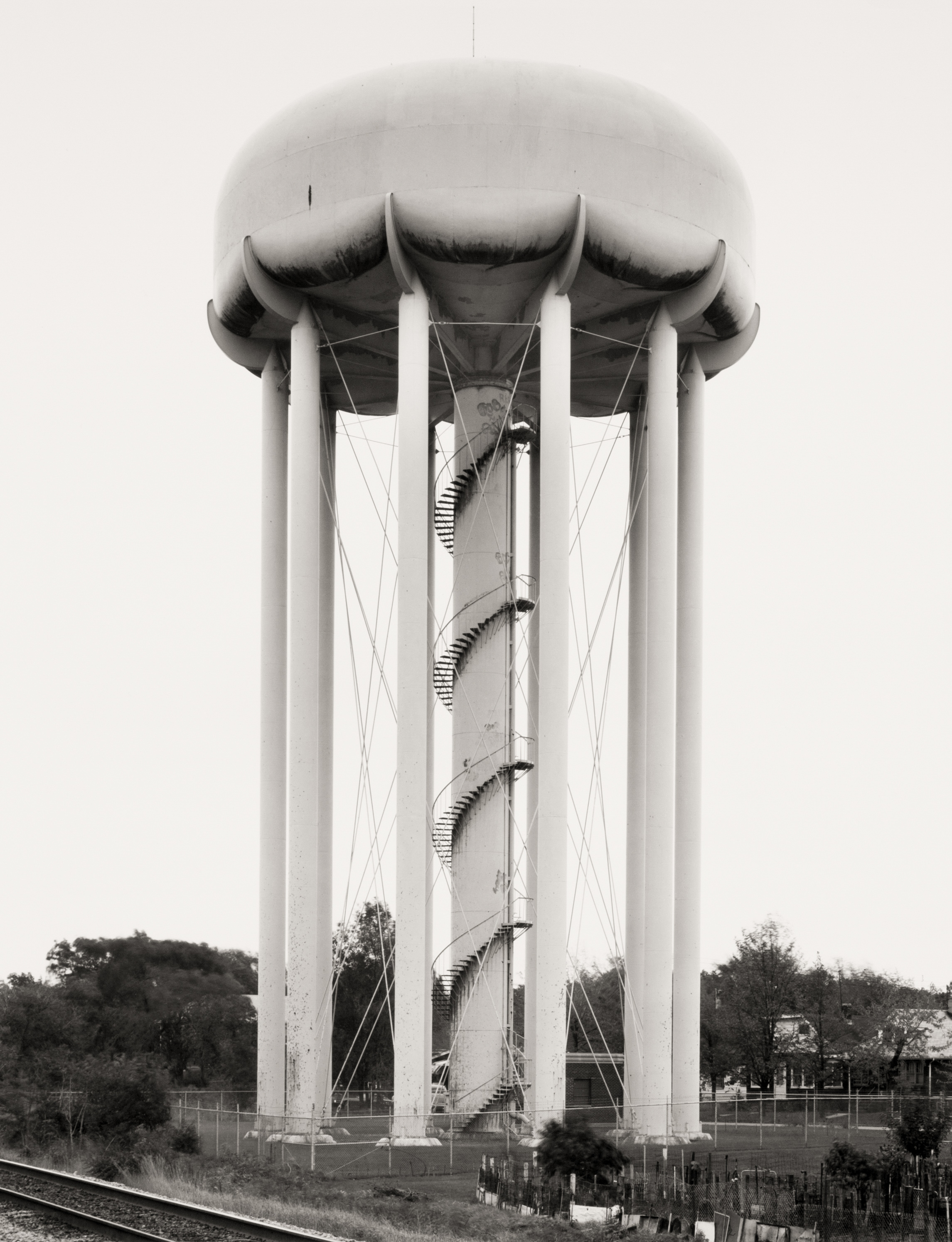 Black and white photograph of a water tower near a railroad track and residential homes