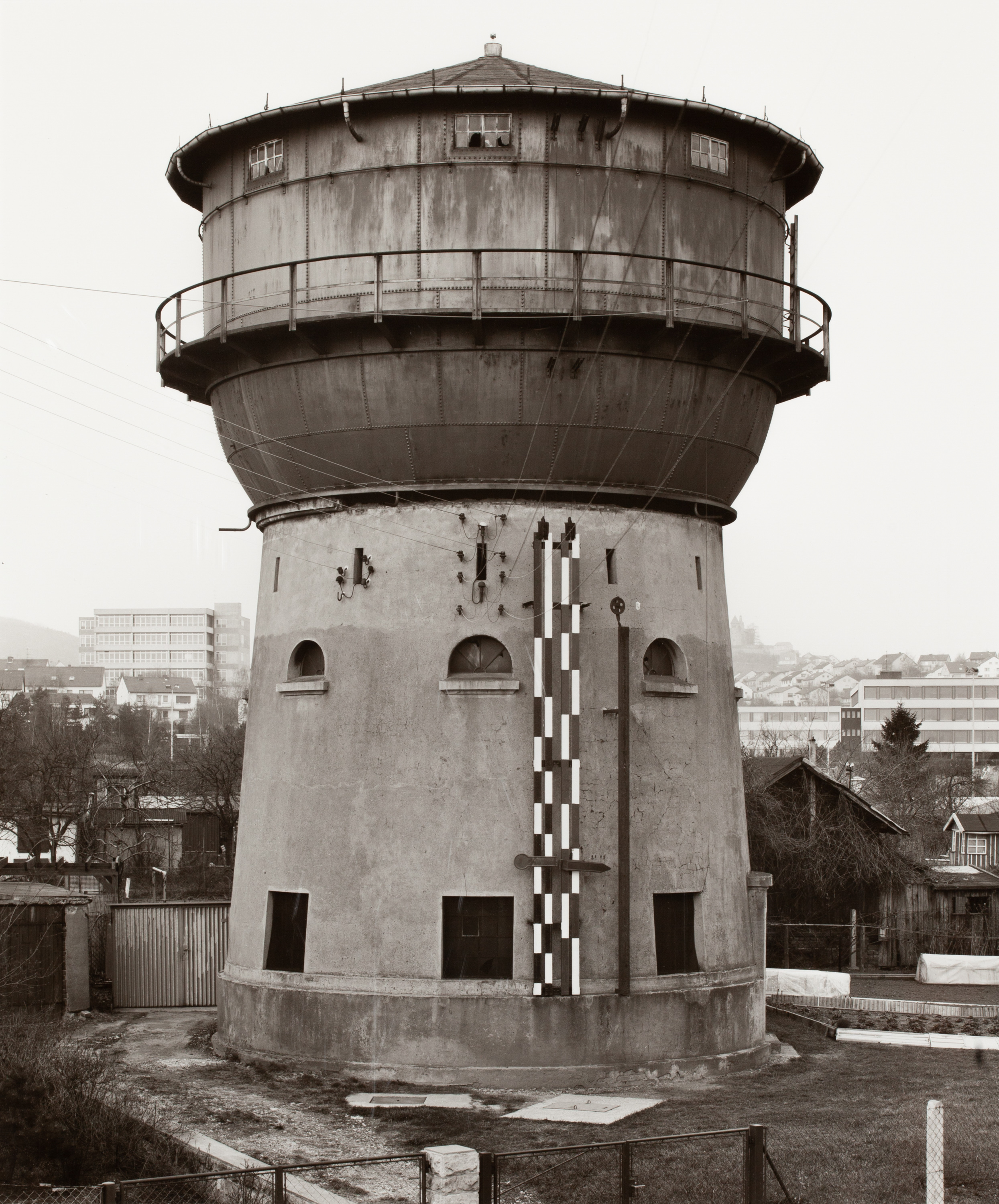Black and white photograph of a water tower among residential homes with larger city on the horizon