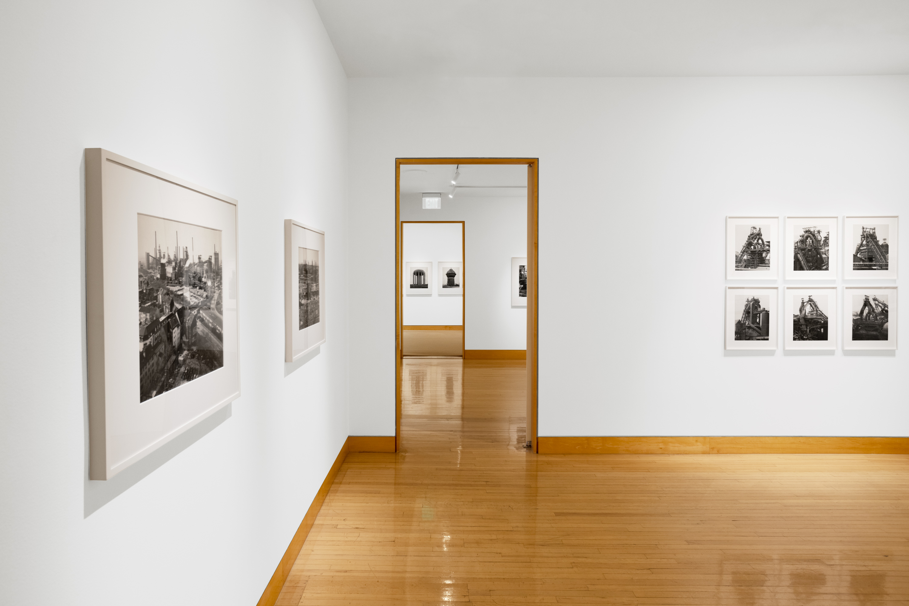 Color image of black and white photographs of industrial buildings and furnaces, framed in white on white gallery walls