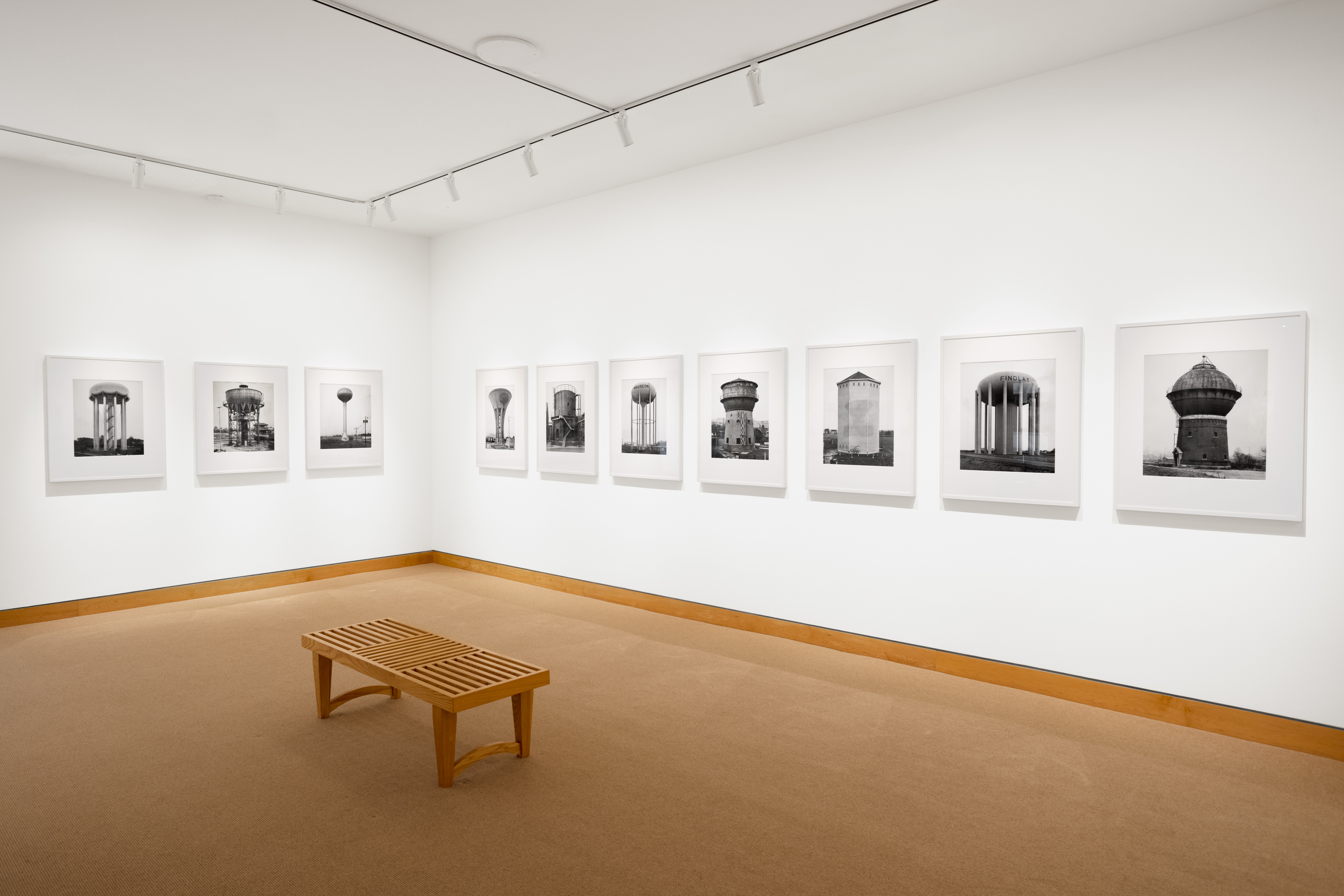Color image of black and white photographs of various water towers framed in white on white gallery walls