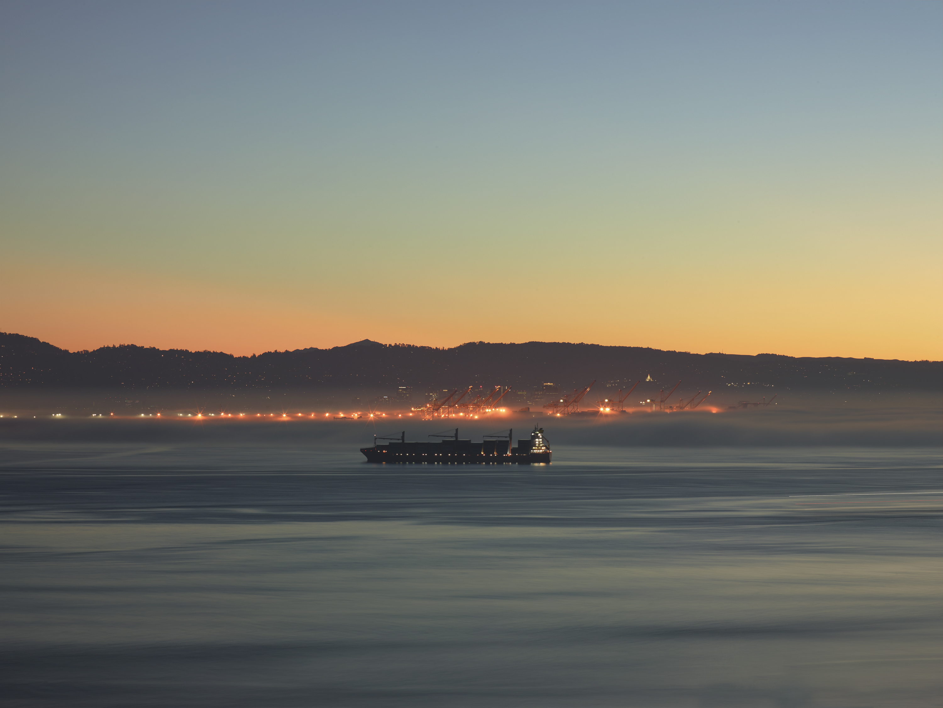 Color photograph of a cargo ship moving across the water around sunrise