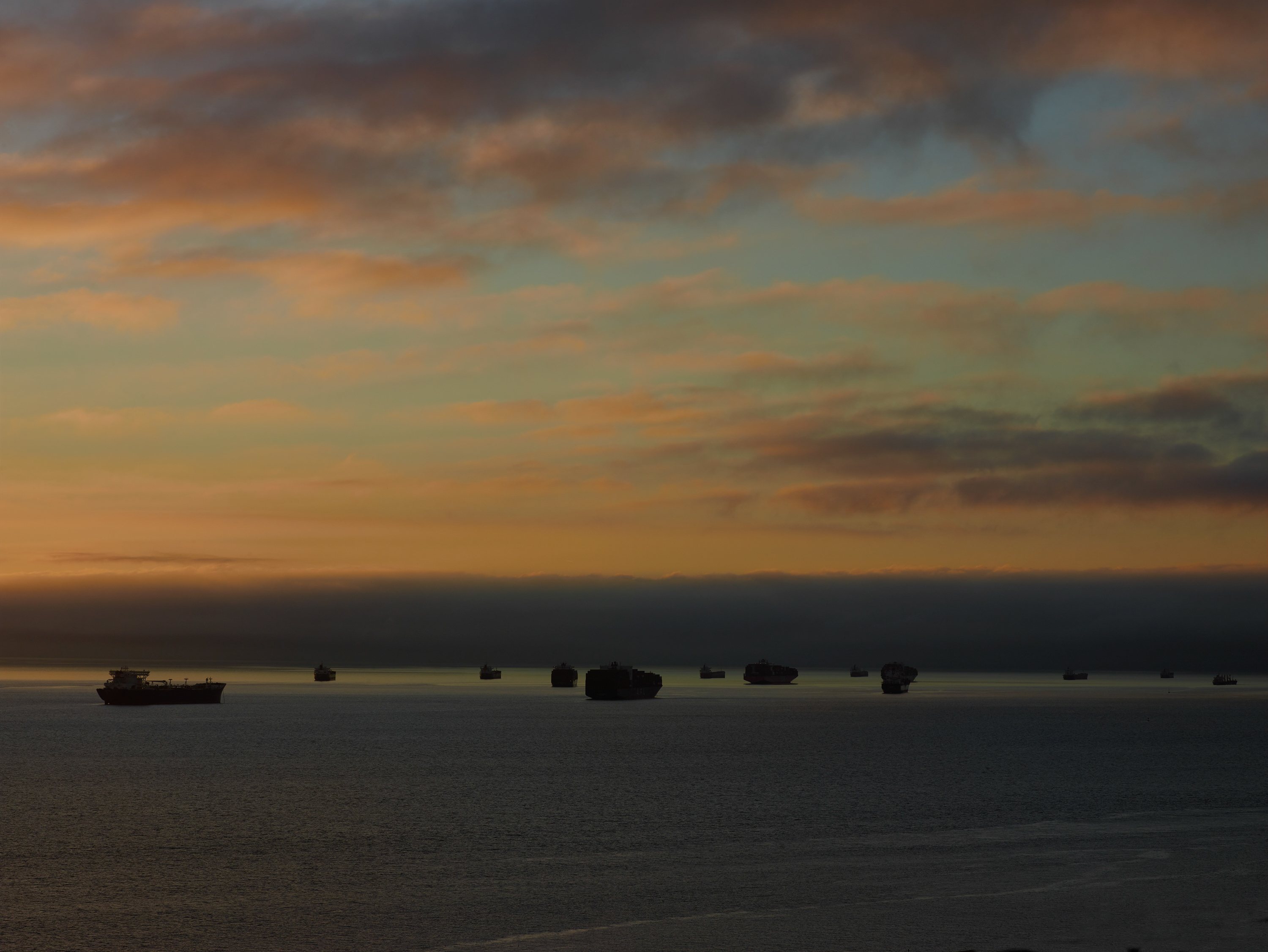 Color photograph of cargo ships on the water with a large cloud of fog looming on horizon