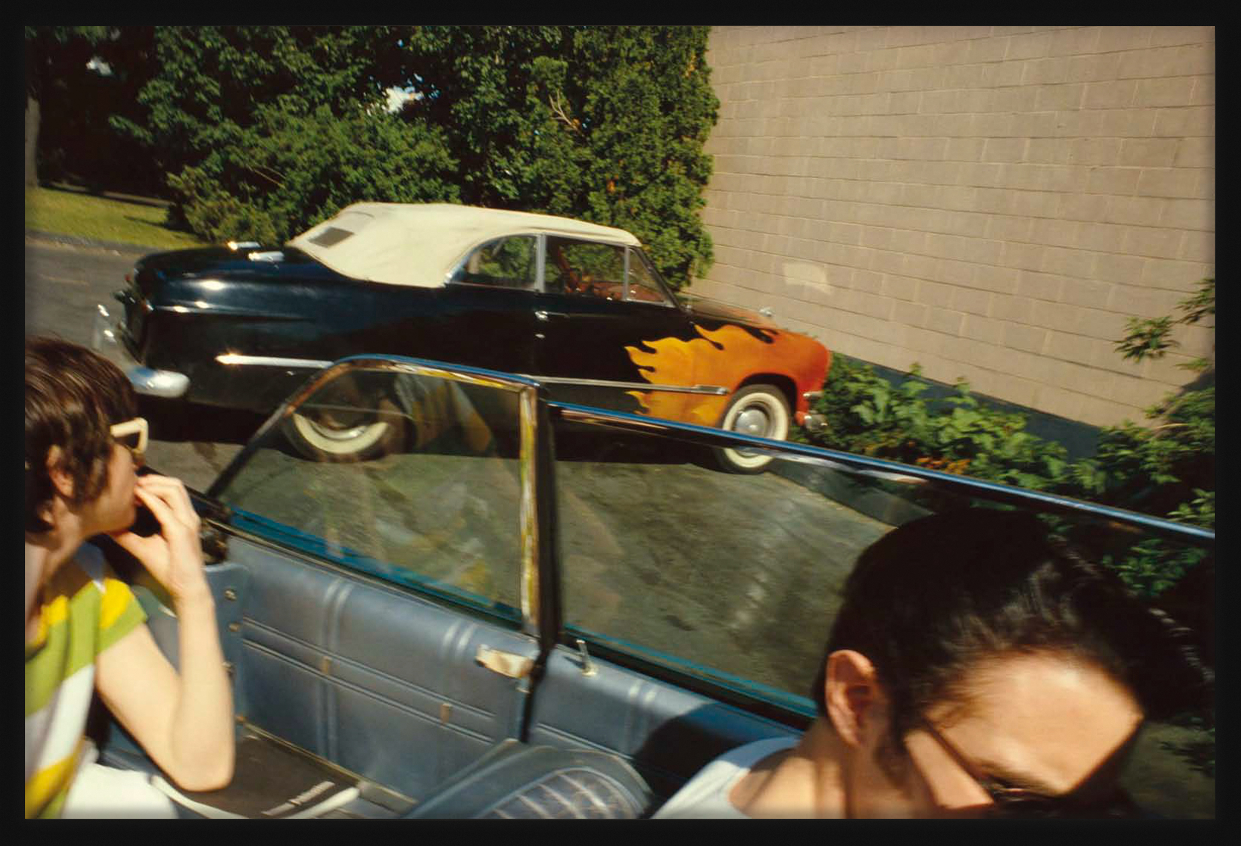Color photograph of a convertible car with painted flames taken from another convertible car framed in black