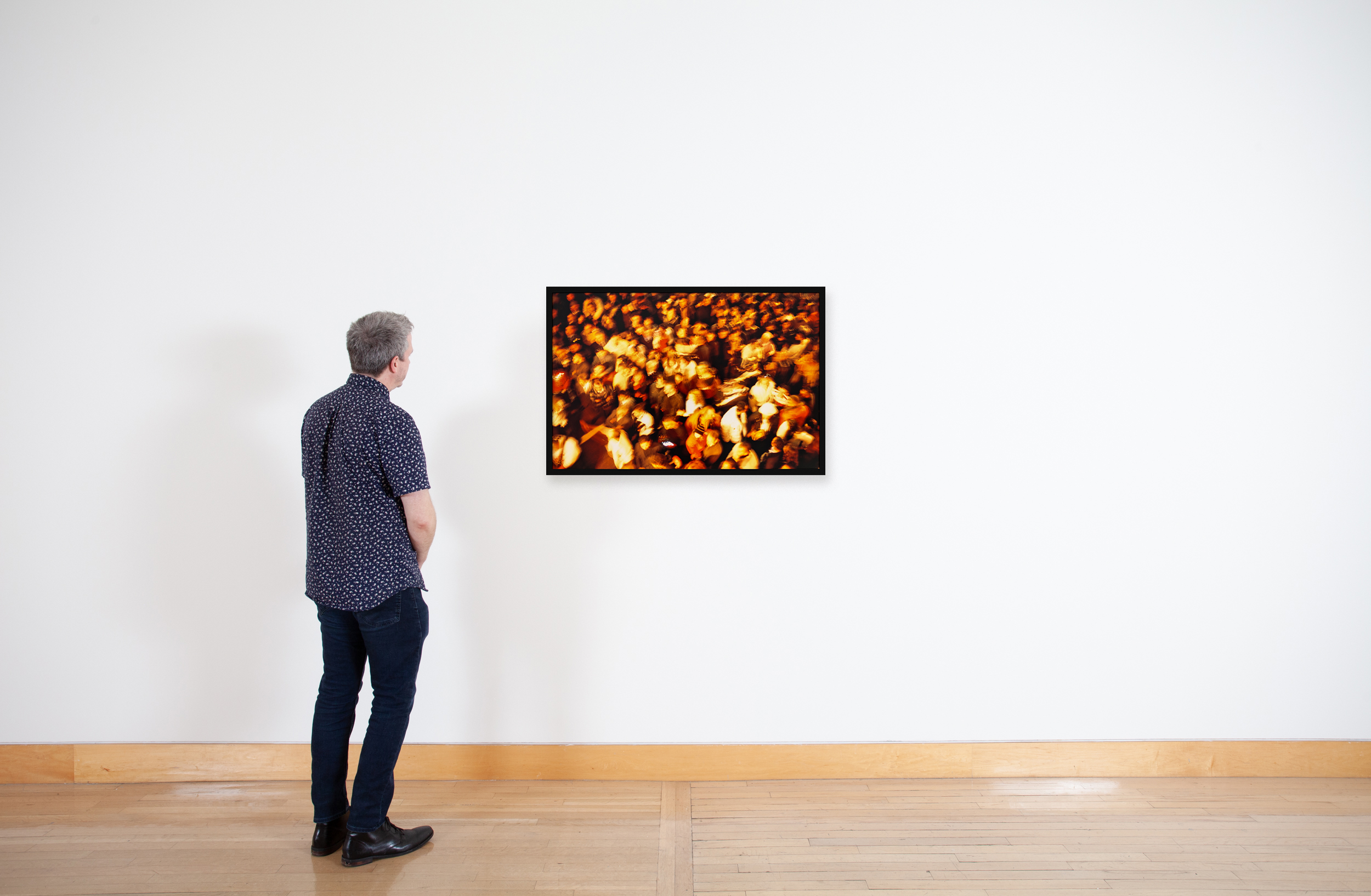 Color image of a color photograph of depicting a warm colored crowd of people seen from above with motion blur on a white gallery wall