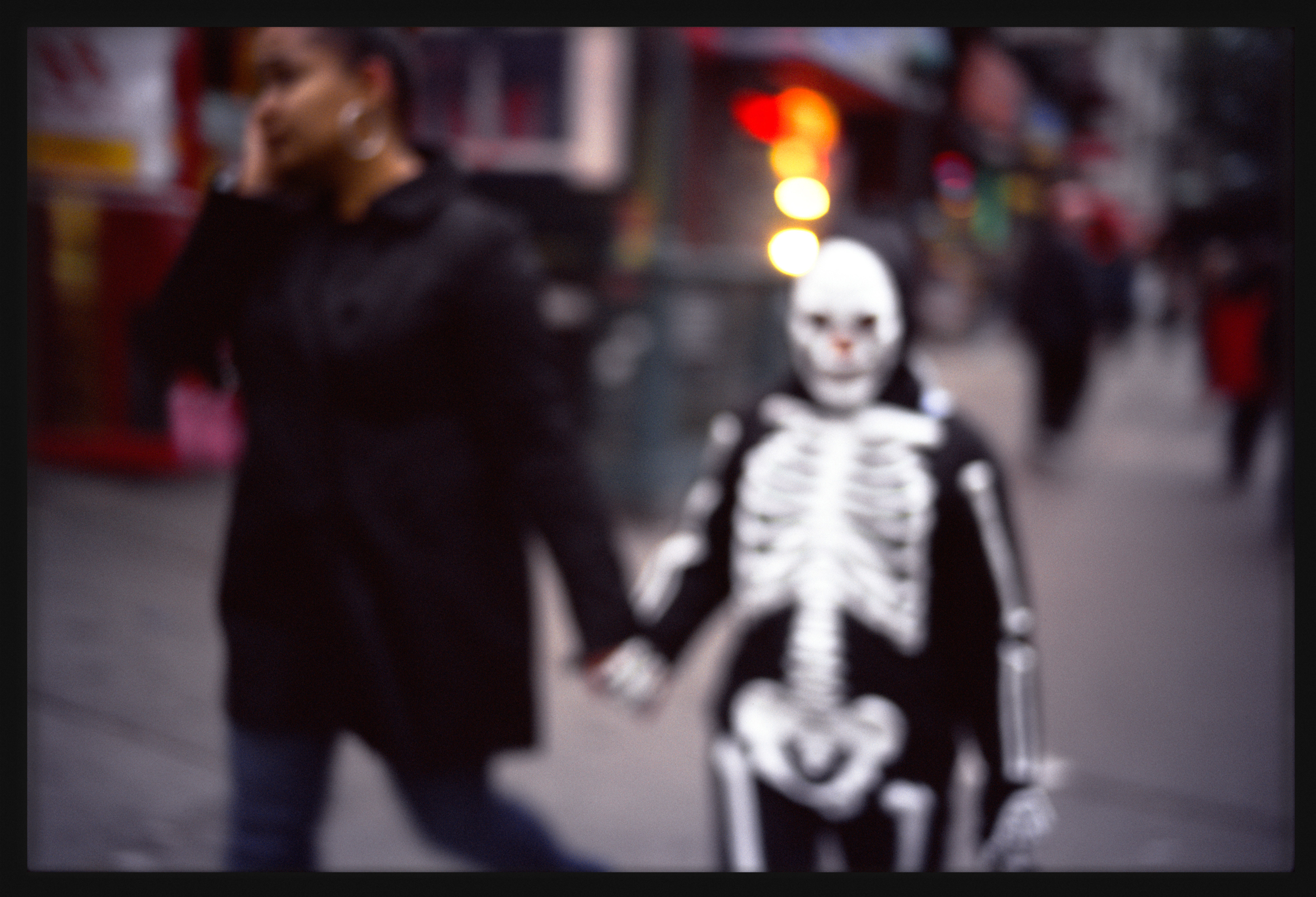 Color photograph of a child in a skeleton costume holding hands with another figure on street framed in black