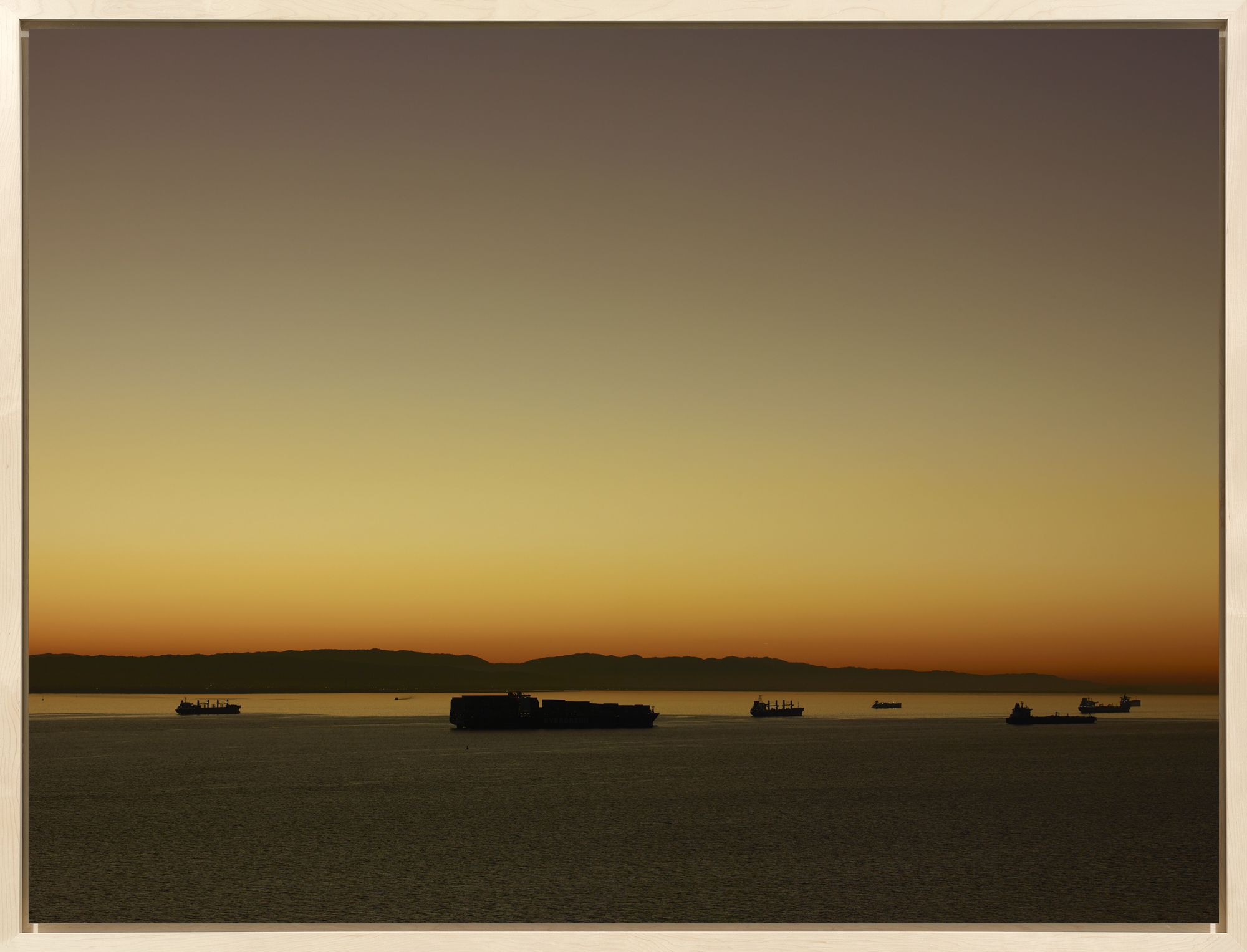 Color photograph of cargo ships moving across the water at around sunrise framed in a bleached wood frame