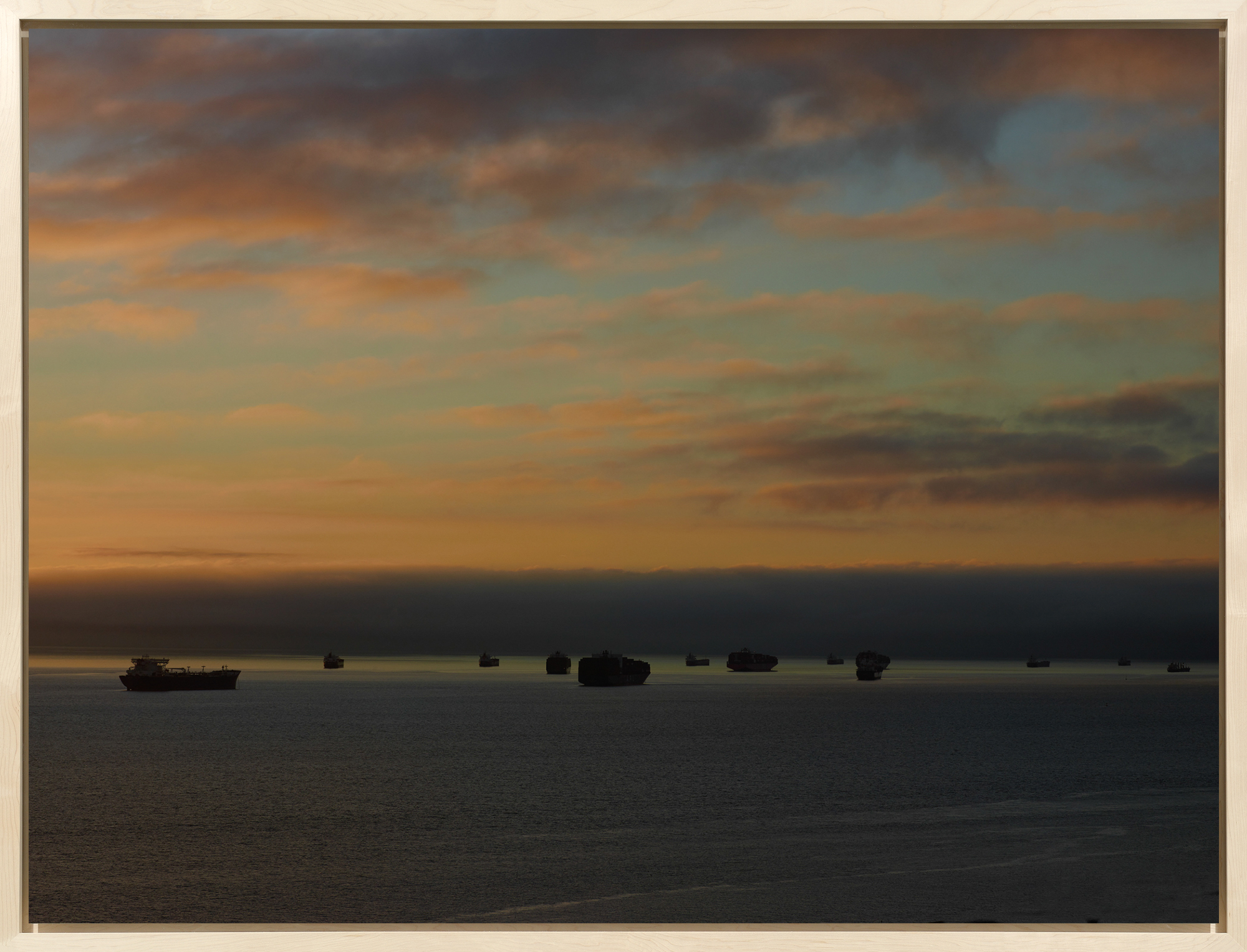 Color photograph of several cargo ships on the horizon during sunrise with pinkish clouds