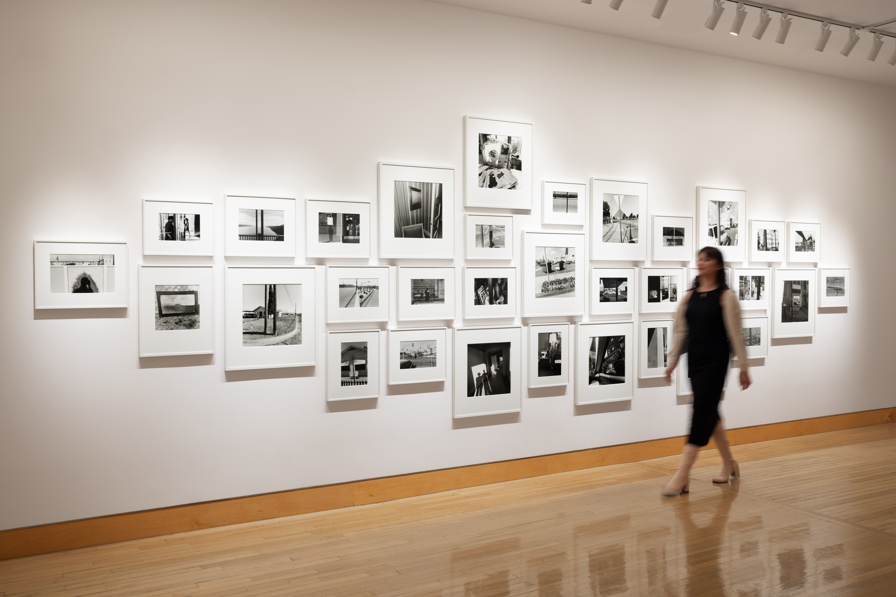 Color image of a gallery exhibiting black and white photographs of various sizes on white gallery wall with blurred figure walking