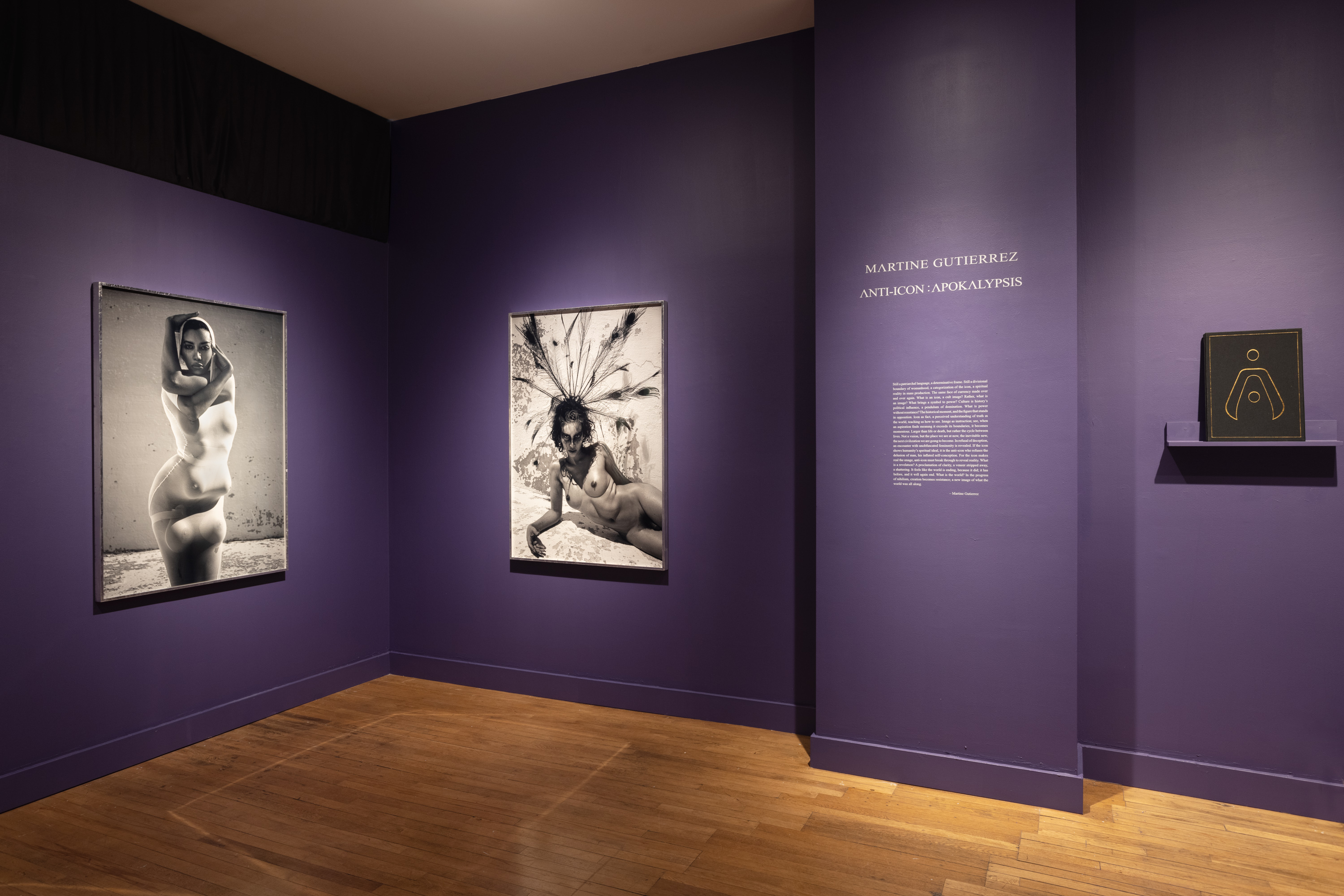 Color image of exhibition with two framed black and white photographs on purple walls with book and exhibition text