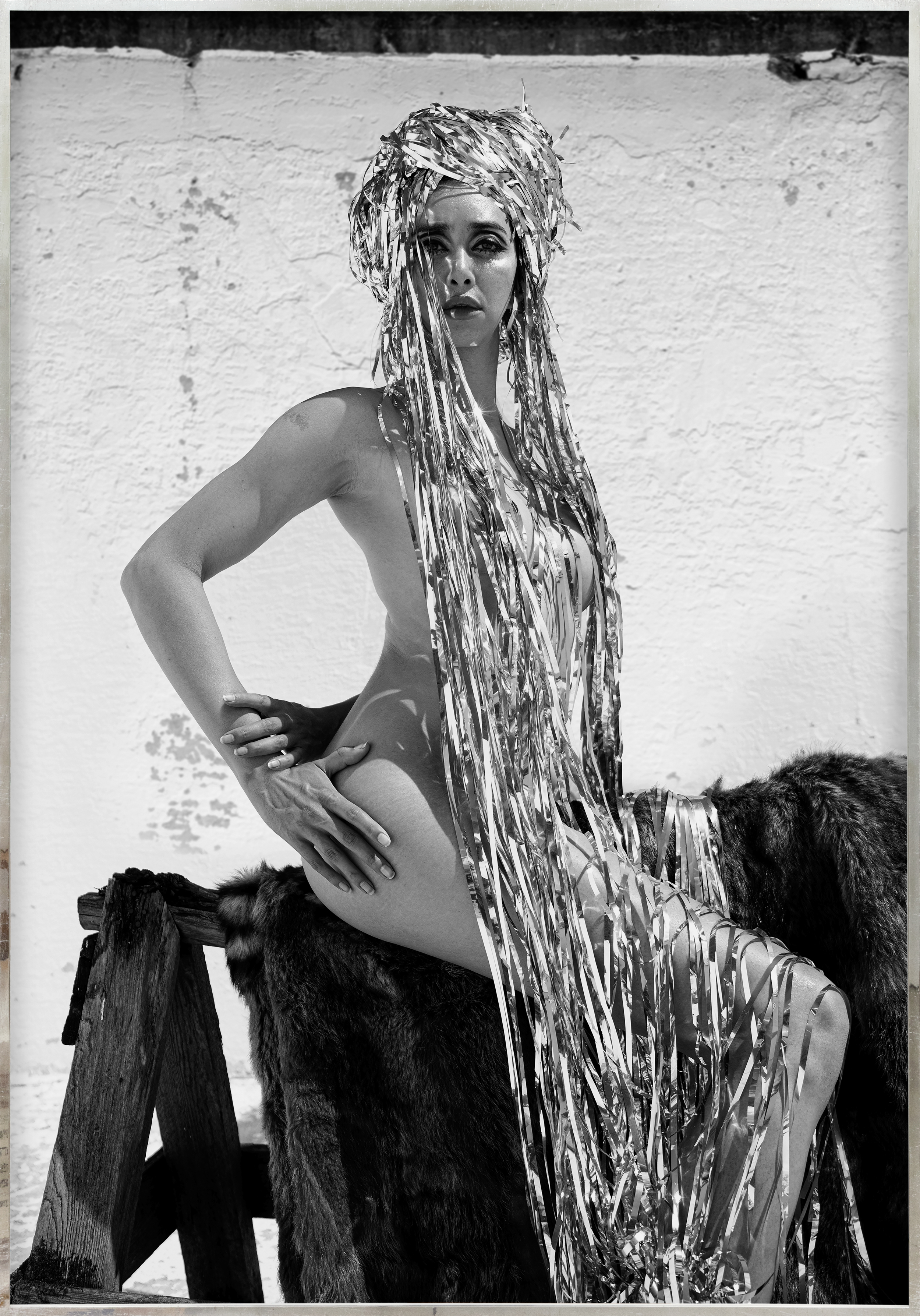 Color image of black and white photograph of a seated figure dressed in a shiny metallic head dress cascading down covering their body framed in a distressed metal